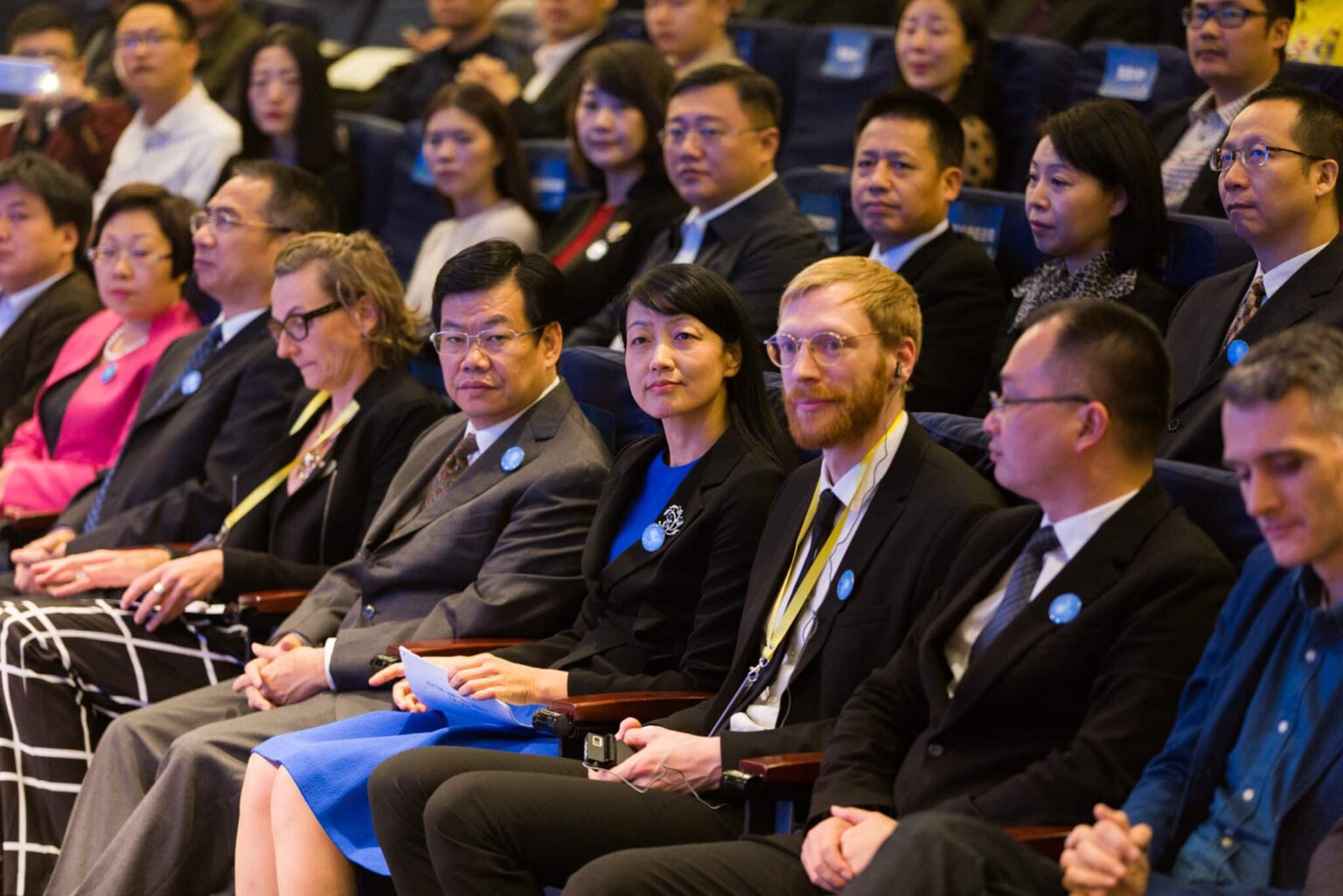 1202xt2080-11 - Deutsche Manufakturenstrasse Personen in formeller Kleidung sitzen in einem Auditorium, einige tragen blaue Anstecker, während sie auf eine Präsentat...