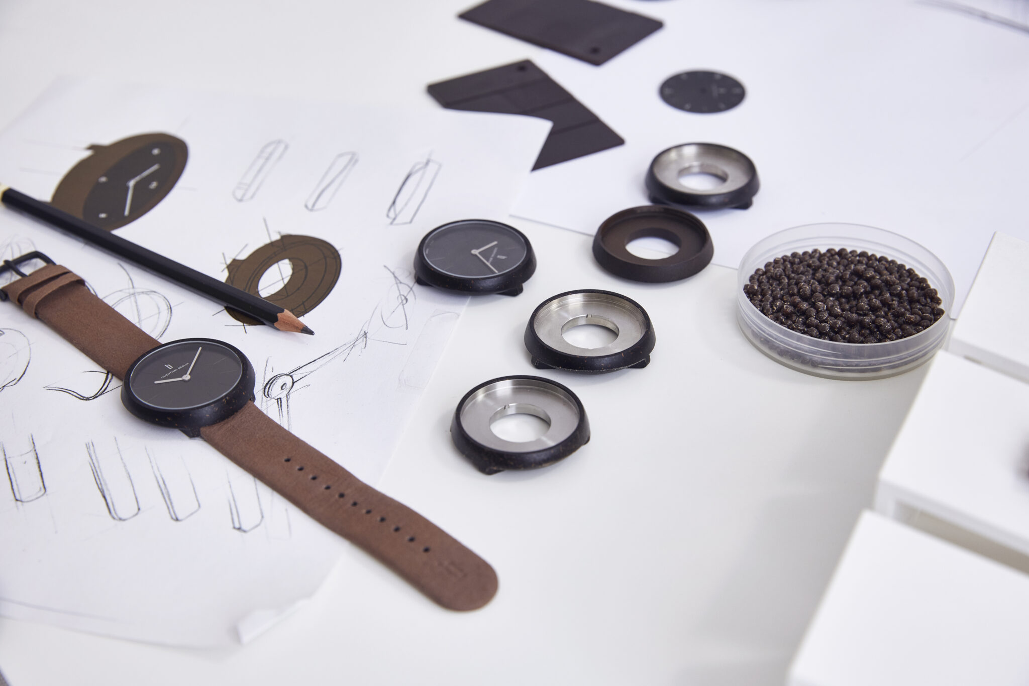 Uhrendesign-Entwürfe mit Skizzen, Uhrengehäusen und einem Armband auf einem weißen Tisch.