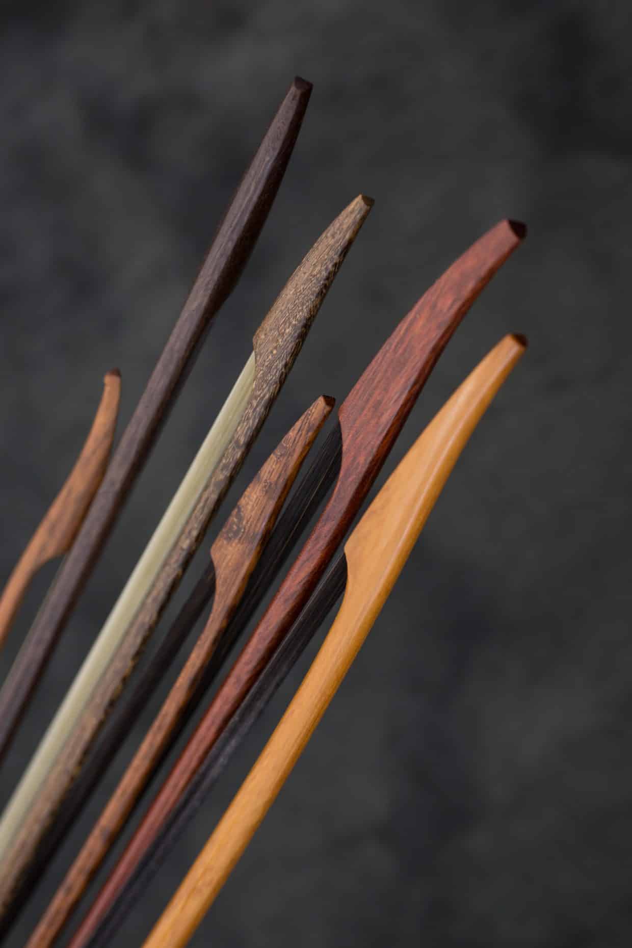 Bogenbuendel1-2484 Kopie - Deutsche Manufakturenstrasse Sechs verschiedene Geigenbogenstangen in unterschiedlichen Holzarten und Farben vor einem dunklen Hintergrund.