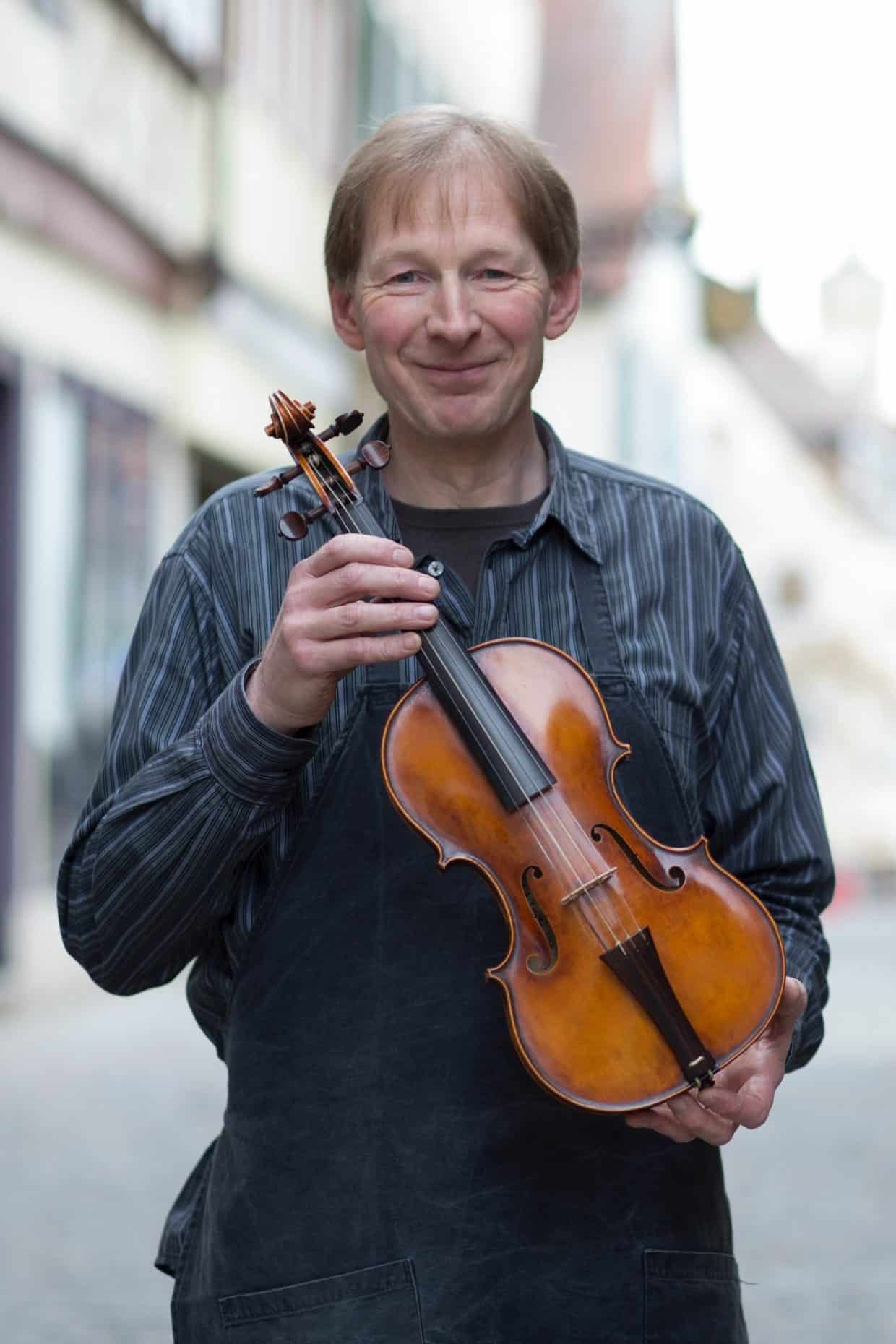 Geigenbauer-5014 Kopie - Deutsche Manufakturenstrasse Geigenbauer hält eine Geige in der Hand, lächelt und steht in einer historischen Straße.