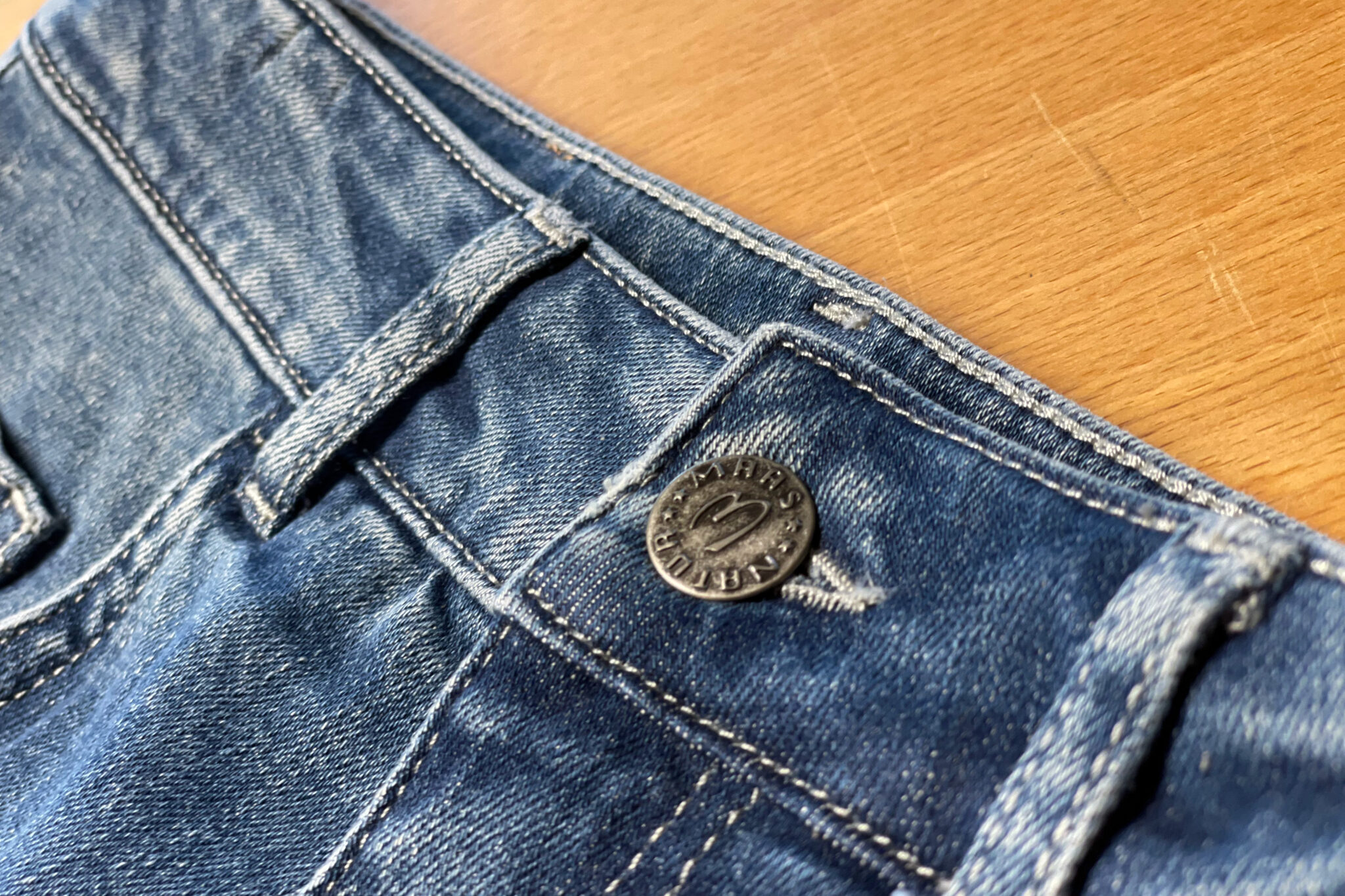 Detailaufnahme einer Jeans mit sichtbarem Knopf und Nähte auf einem Holzuntergrund.