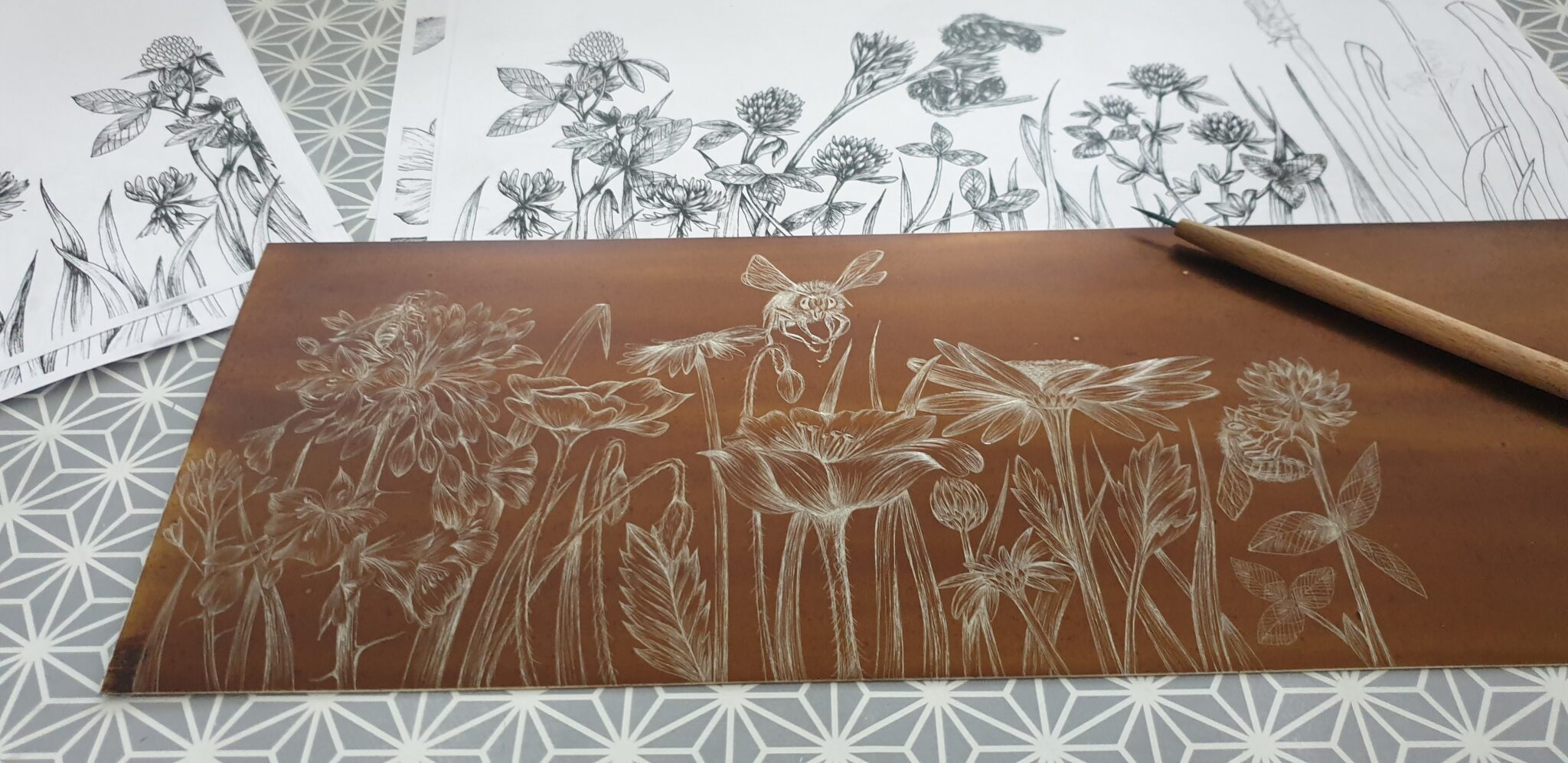 Detailaufnahme einer Kupferplatte mit eingravierten Blumen und einer Biene, umgeben von Skizzen auf Papier.