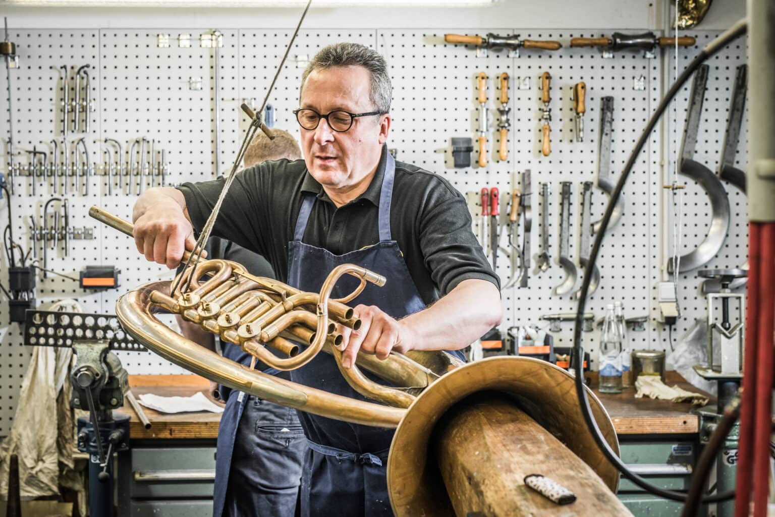 Handwerker in einer Werkstatt, der eine große Messing-Trompete bearbeitet, umgeben von Werkzeugen und Materialien.