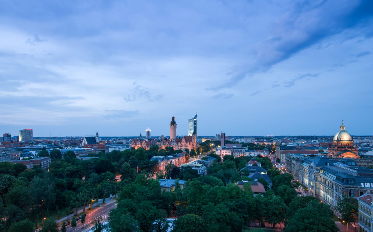 Skyline von Leipzig bei Dämmerung mit dem Neuen Rathaus und Hochhäusern, umgeben von Bäumen und Stadtlichtern.