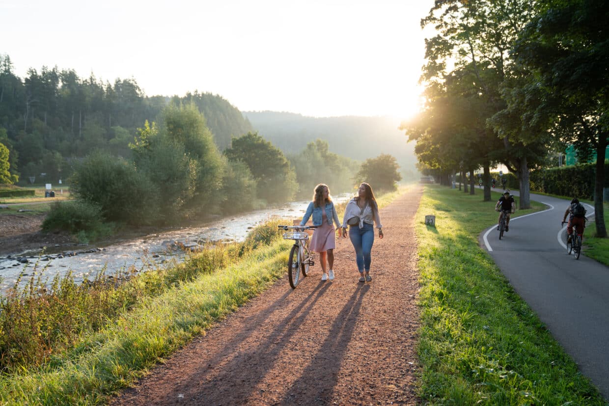 Zwei Frauen spazieren entlang eines schmalen Weges neben einem Fluss, umgeben von Bäumen, bei Abendstimmung.