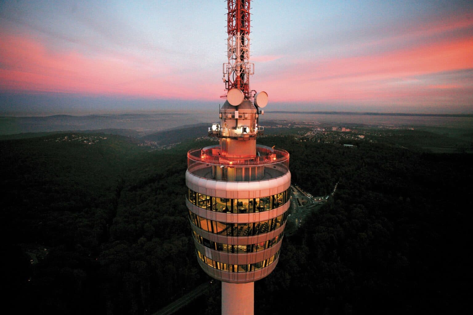 - Deutsche Manufakturenstrasse TV-Turm Stuttgart bei Sonnenuntergang, beleuchtet mit orangefarbenem Licht, umgeben von bewaldeten Hügeln und einem farbe...