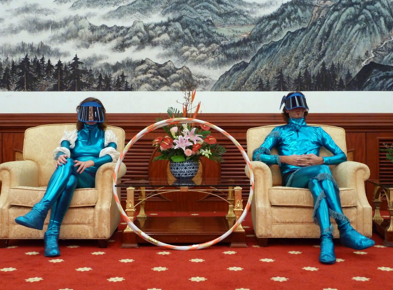 - Deutsche Manufakturenstrasse Zwei Personen in blauen, futuristischen Anzügen sitzen auf beigen Sofas, umgeben von einer Pflanzenarrangement und einer ...