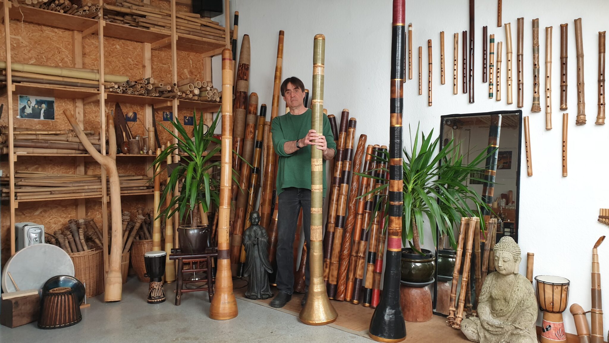 Michael-Marahrens-Didgeridoos_gross - Deutsche Manufakturenstrasse Mann steht in einem Raum voller Didgeridoos, umgeben von Pflanzen und Musikinstrumenten.