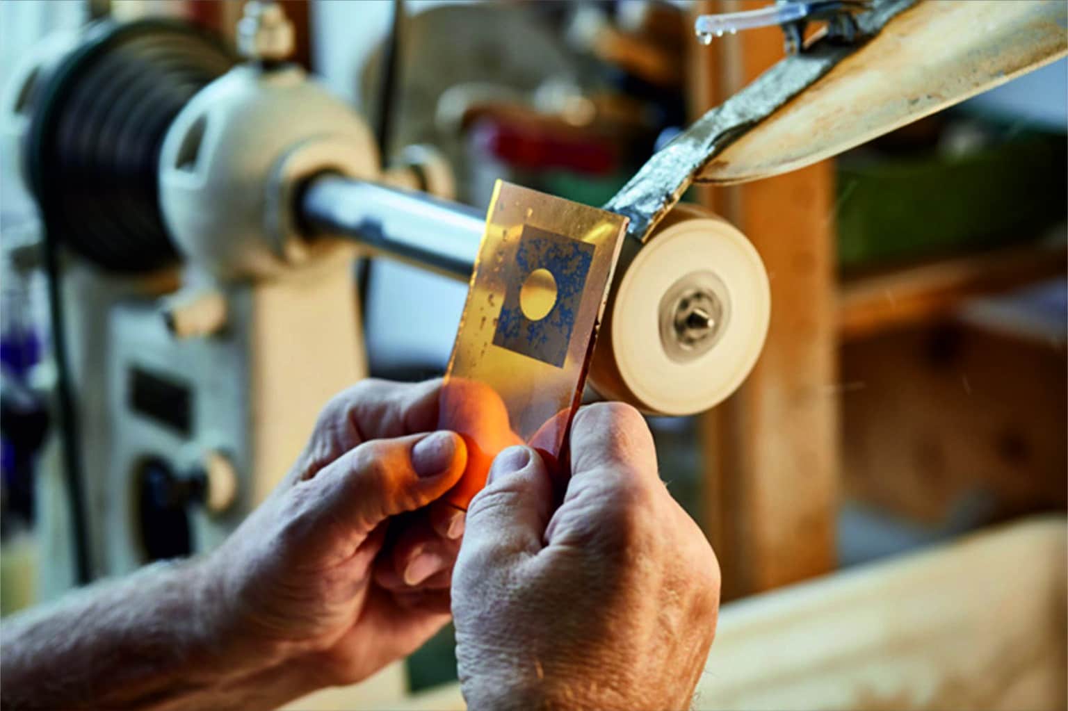 - Deutsche Manufakturenstrasse Hände halten ein goldenes Glasstück vor einer Schleifmaschine in einer Werkstatt.