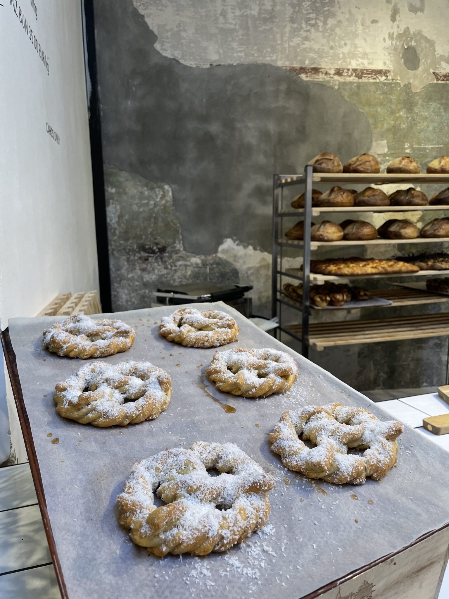 Zwei Bleche mit Zimtgebäck und Puderzucker, im Hintergrund frisch gebackenes Brot in einem modernen Bäckerei-Interieur.