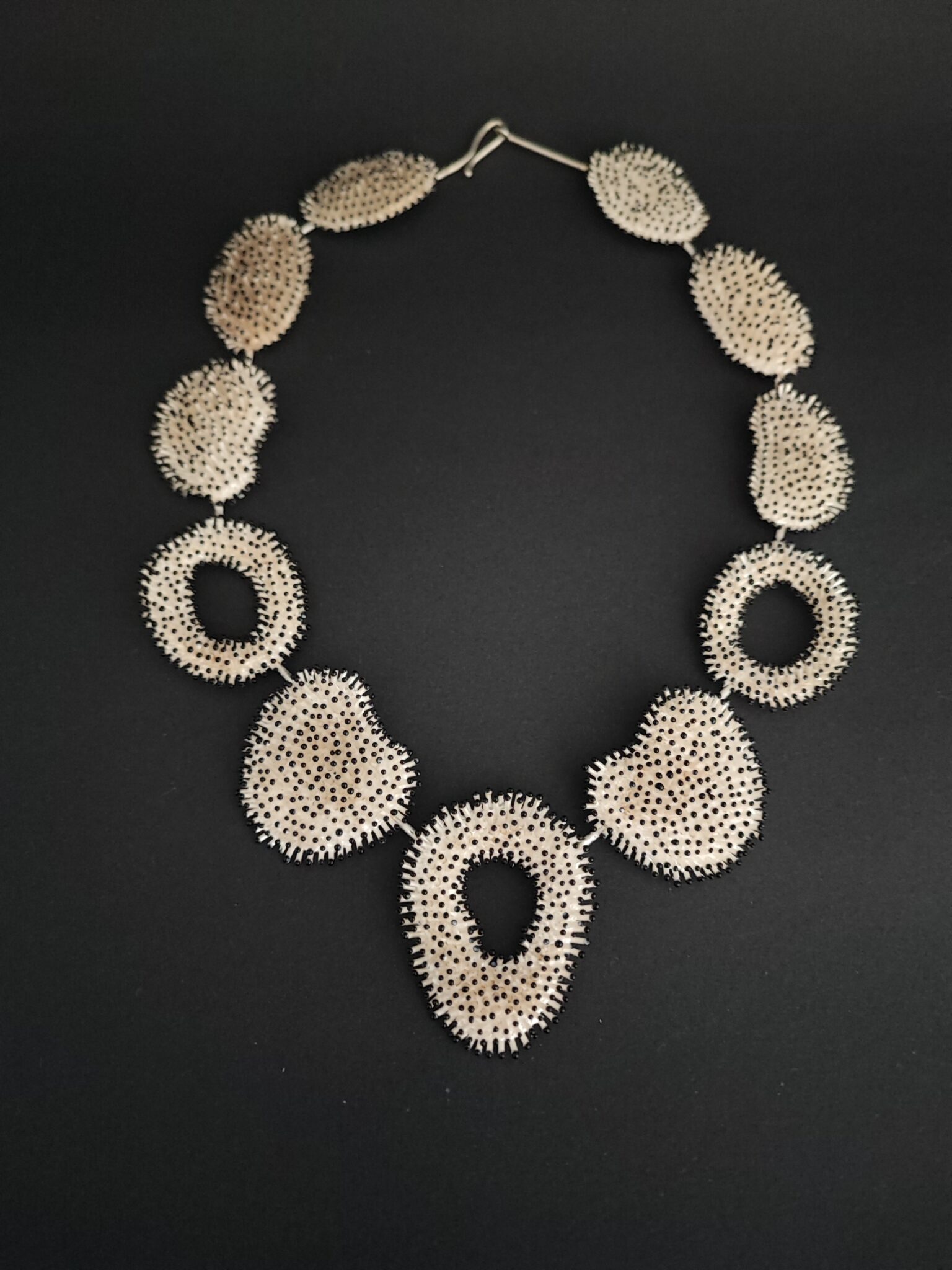 1674404754898 - Deutsche Manufakturenstrasse Kunstvoll gestaltete Halskette aus hellen, ovalen Elementen mit schwarzen Punkten auf schwarzem Hintergrund.