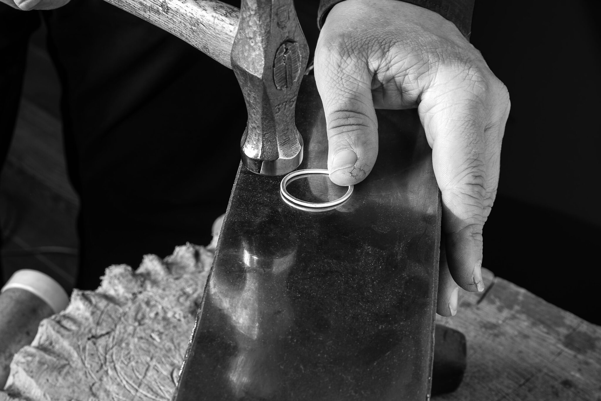 Hand mit Hammer, die einen silbernen Ring auf einer Metallplatte bearbeitet, in Schwarz-Weiß.