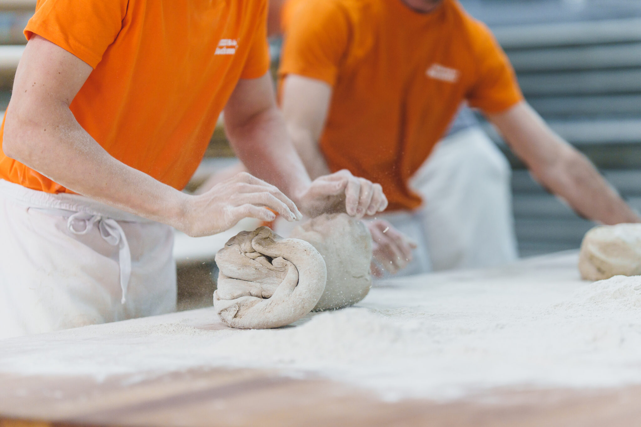 Bäcker Bachmeier - Deutsche Manufakturenstrasse Zwei Bäcker in orangefarbenen T-Shirts kneten Teig auf einer mit Mehl bestäubten Arbeitsfläche.