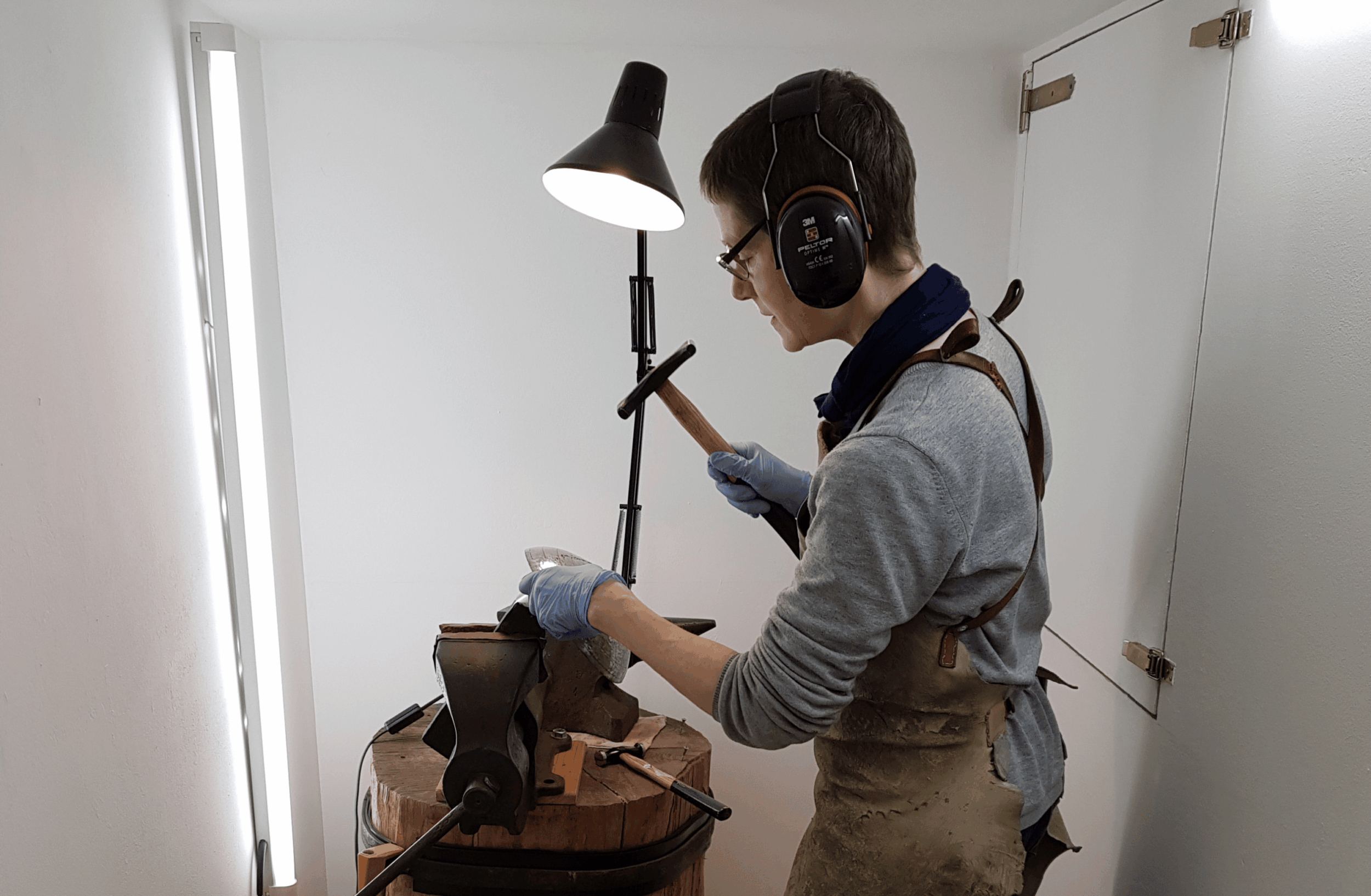 Person mit Kopfhörern und Schutzhandschuhen arbeitet an einem Werkstück an einer Werkbank, beleuchtet von einer Lampe.