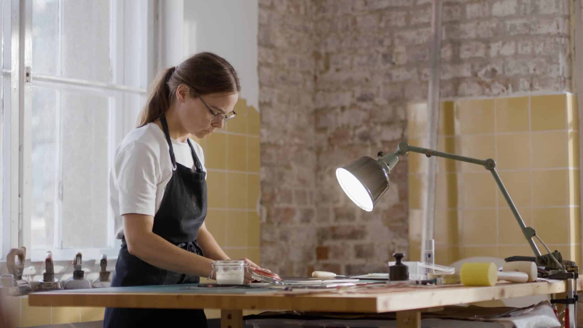 Frau in schwarzer Schürze arbeitet konzentriert an einem Tisch in einer hellen Werkstatt mit Backsteinwand.