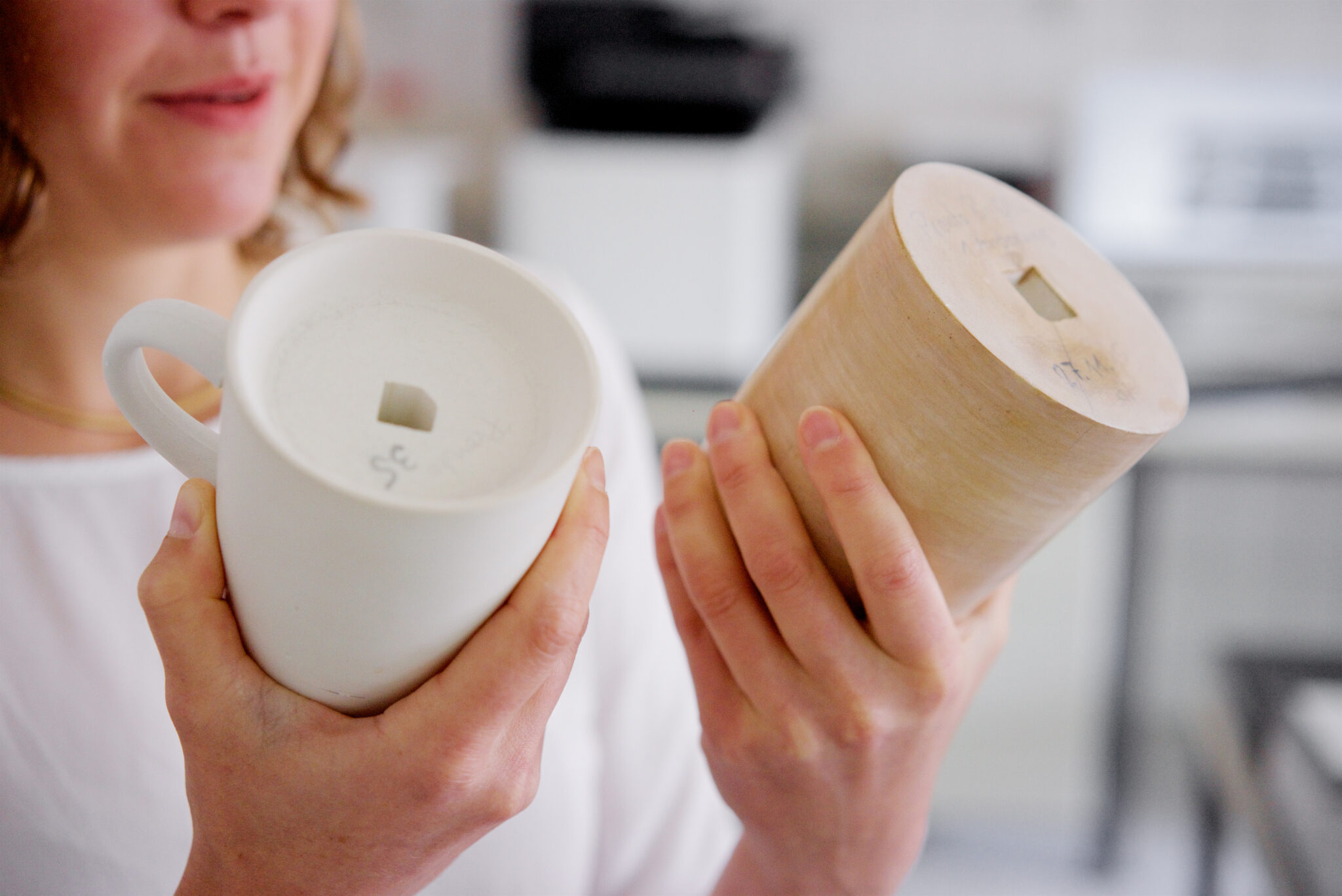 - Deutsche Manufakturenstrasse Frau hält zwei Porzellanobjekte, einen weißen Becher und einen zylindrischen Rohling, in einer Werkstatt der Porzellanma...