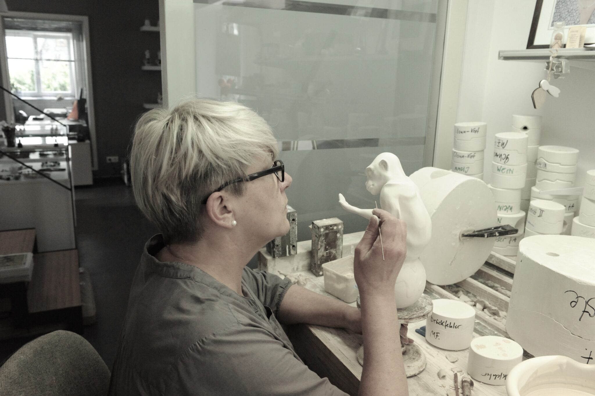 - Deutsche Manufakturenstrasse Künstlerin arbeitet an einer weißen Skulptur eines Affen in einem Atelier, umgeben von Töpfermaterialien.