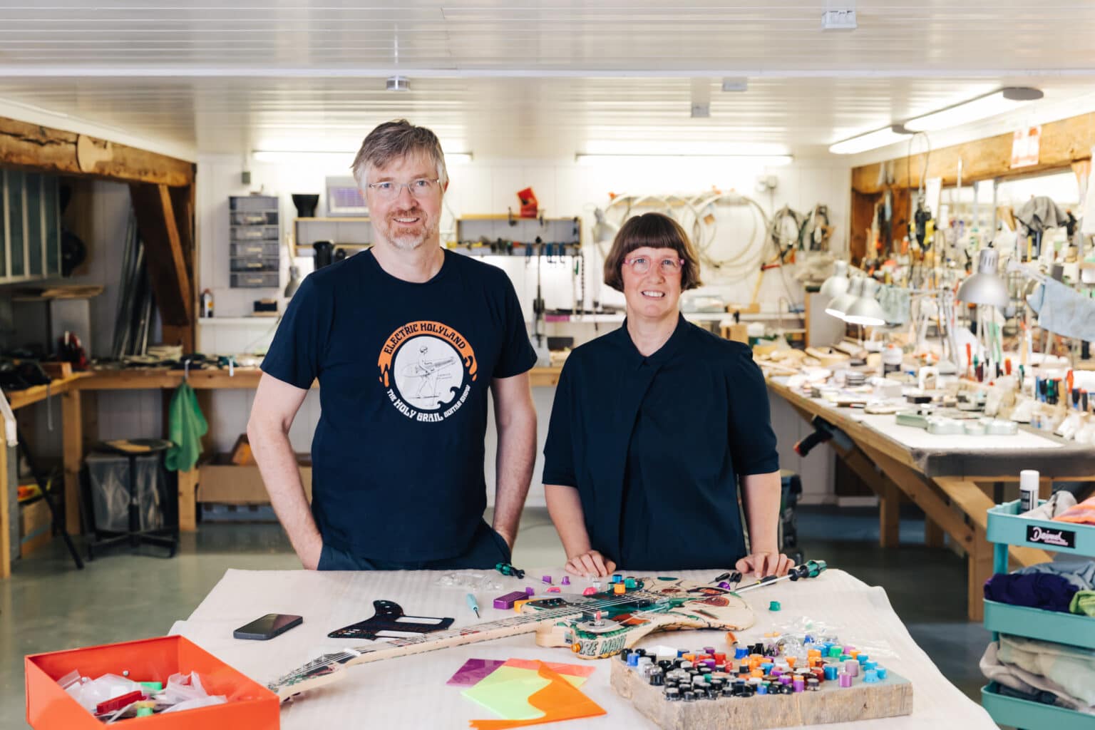 2023_07_Deimel_Guitarworks-1424 - Deutsche Manufakturenstrasse Zwei Personen stehen in einer Werkstatt von Deimel Guitarworks, umgeben von Werkzeugen und Materialien zur Gitarrenherstel...