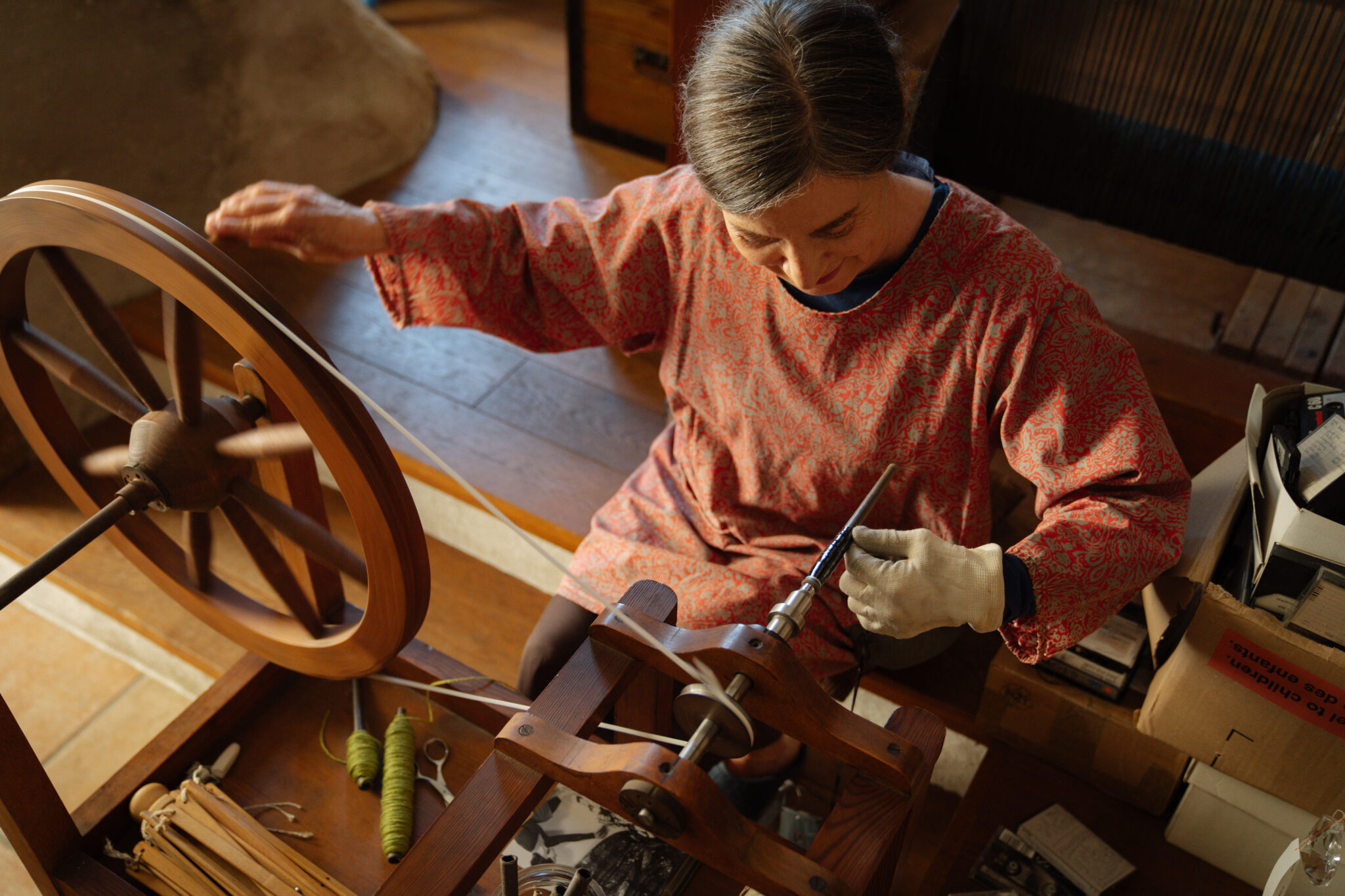 Frau in einem roten Gewand arbeitet an einem Spinnrad in einem hellen Raum mit Holzdetails.