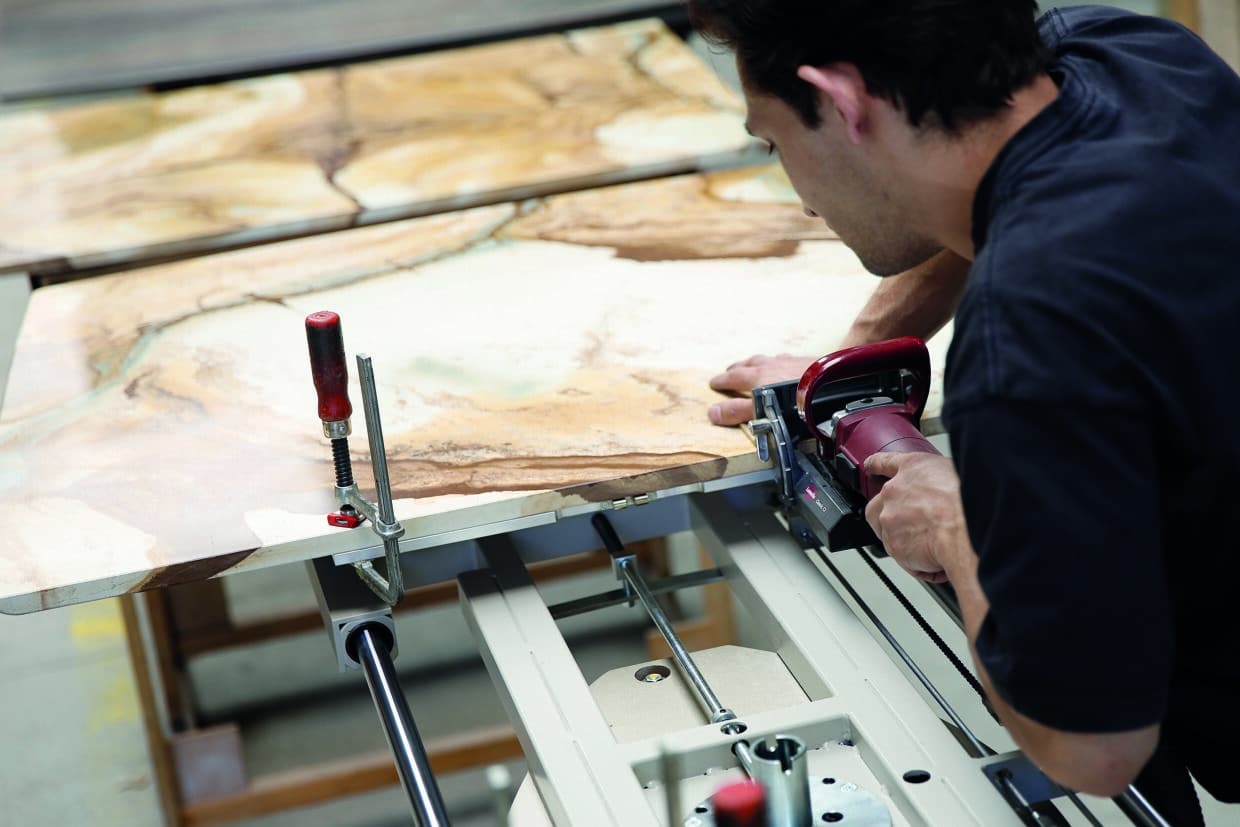 Draenert - Deutsche Manufakturenstrasse Handwerker schneidet mit einer Maschine eine große Platte aus Stein oder Holz, umgeben von Werkbänken.