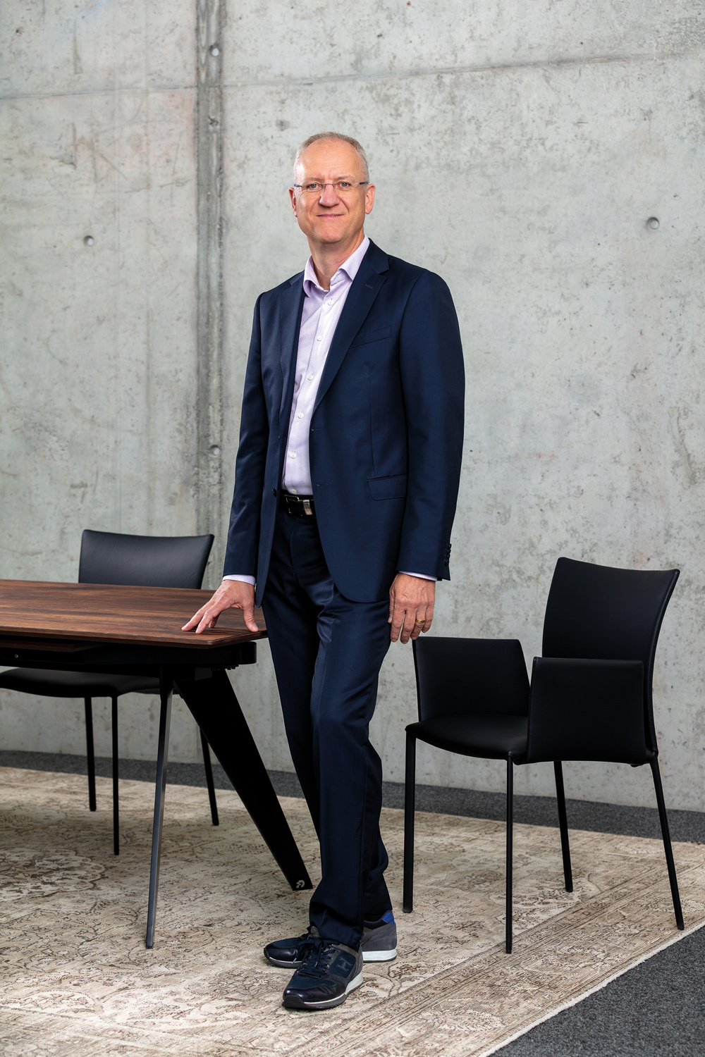 DRAENERT_Portrait-Dr-Patric-Draenert_2023_09_004 - Deutsche Manufakturenstrasse Dr. Patric Draenert steht in einem modernen Büro vor einem Tisch und zwei Stühlen, mit einem neutralen Betonhintergrund.