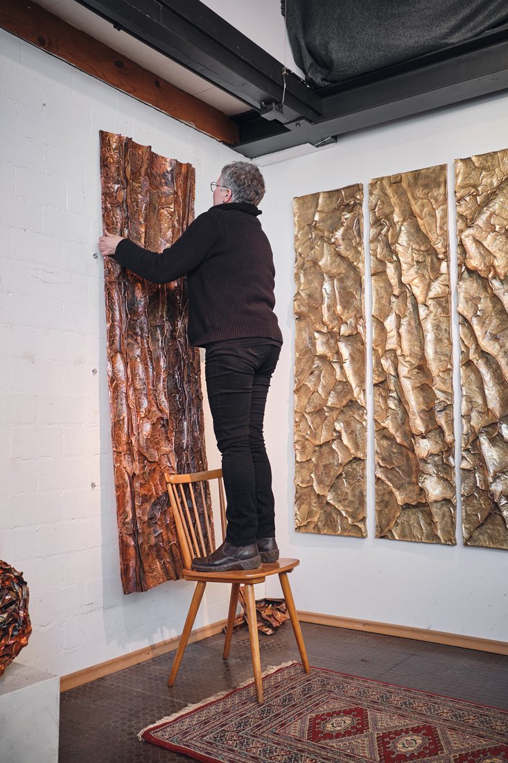 Manuela-Geugelin-2-2 - Deutsche Manufakturenstrasse Künstler hängt eine strukturierte Wandinstallation in einem Atelier auf, während er auf einem Stuhl steht.