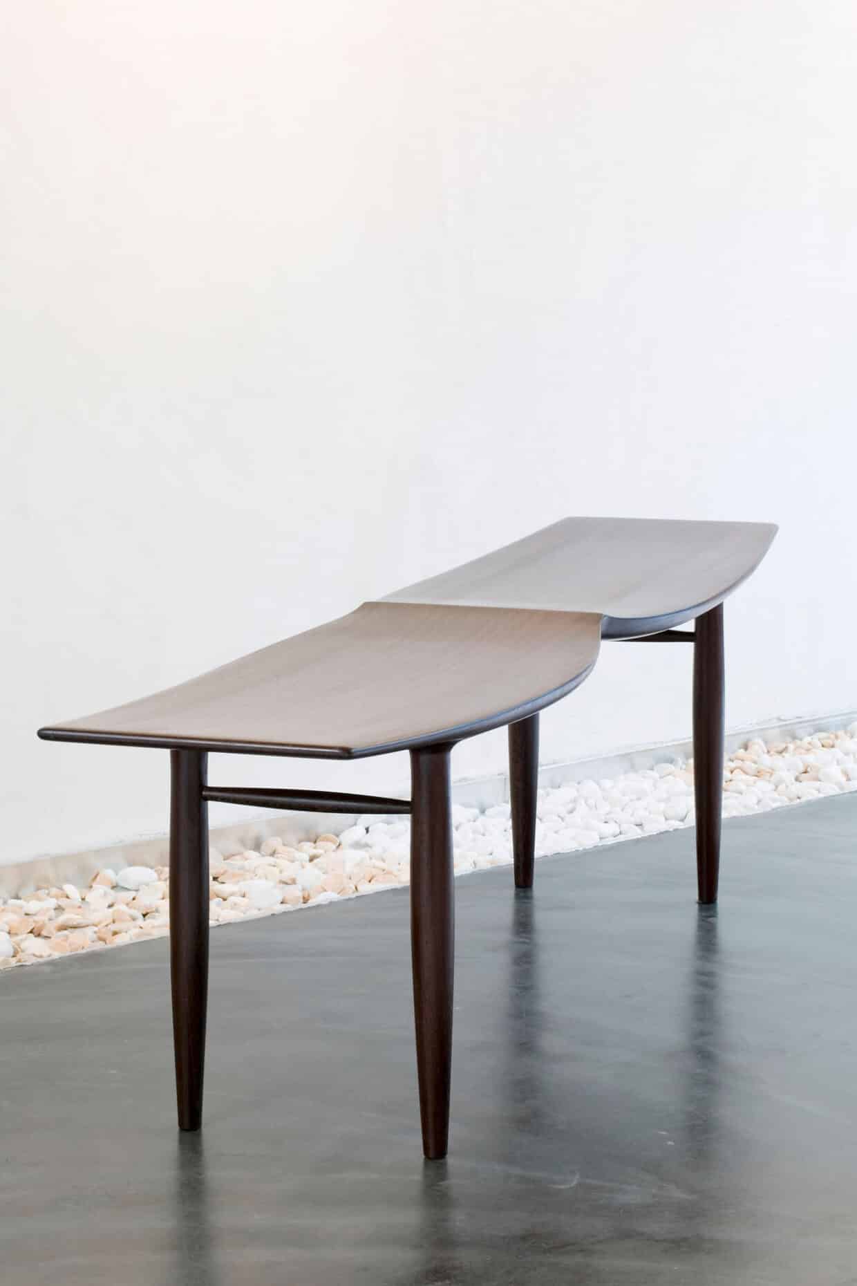 Möbelstücke nr 8 - Deutsche Manufakturenstrasse Sitzbank aus Wengeholz mit geschwungener Oberfläche und schlanken Beinen, vor einer hellen Wand.