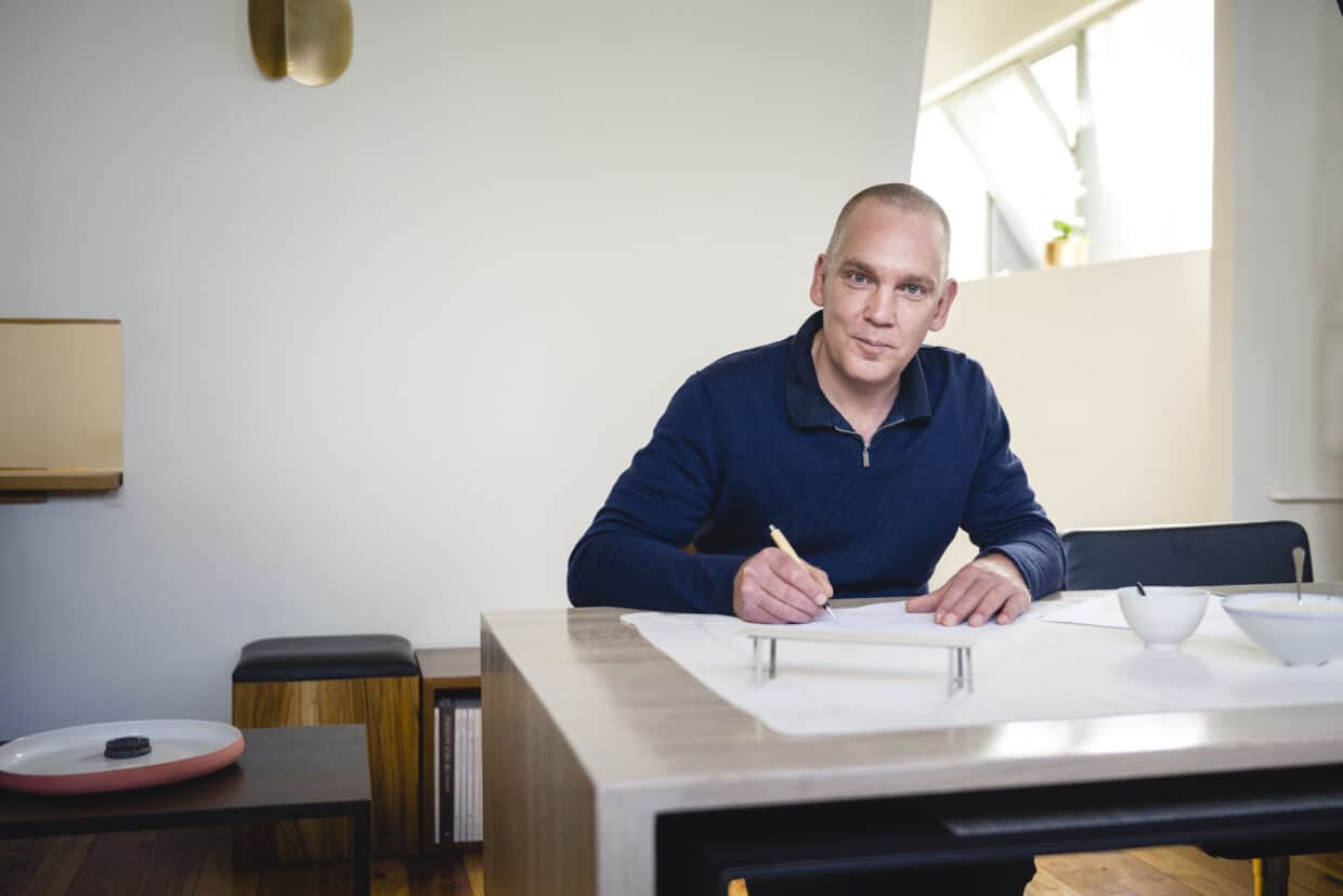 Mann mit kurzen Haaren in blauer Pullover sitzt an einem Tisch und zeichnet auf Papier.