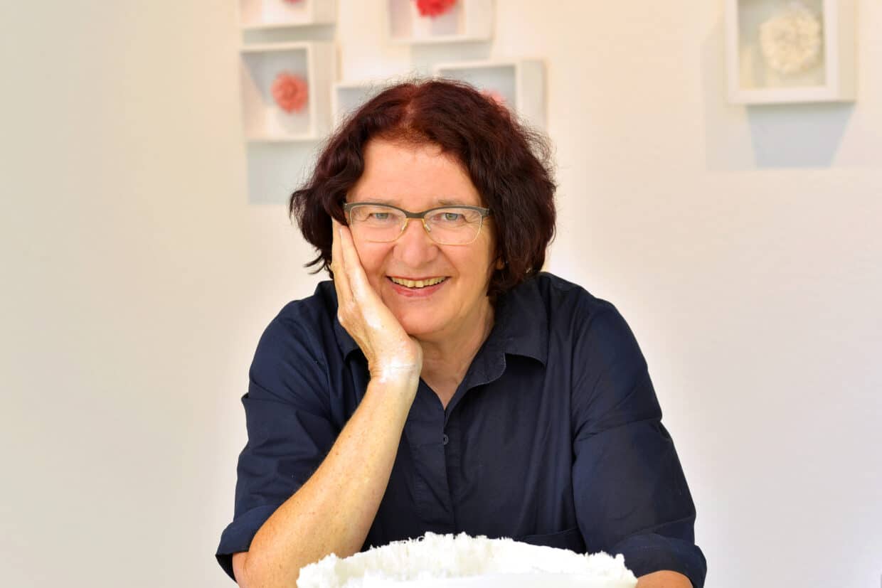 Portrait fotograph klaus ditee - Deutsche Manufakturenstrasse Porträt von Heide Nonnenmacher, lächelnd mit Hand am Gesicht, vor einer hellen Wand mit Kunstwerken.