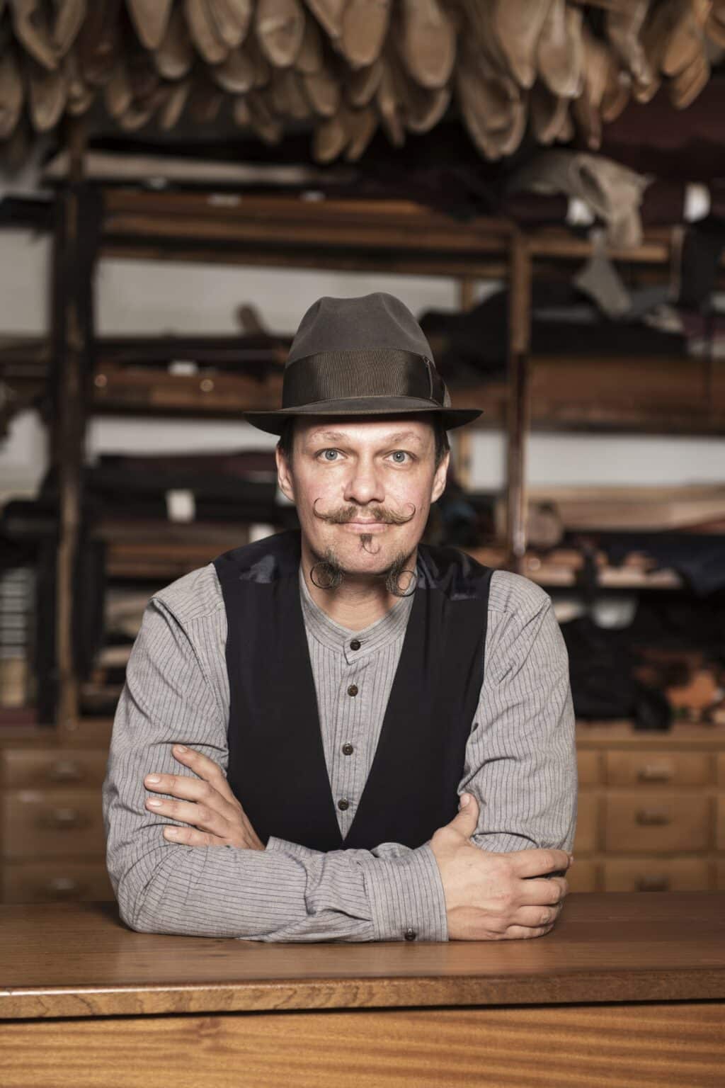 Portrait_neu - Deutsche Manufakturenstrasse Mann mit Hut und Schnurrbart, lächelt am Tisch, umgeben von Schuhmaterialien im Hintergrund.