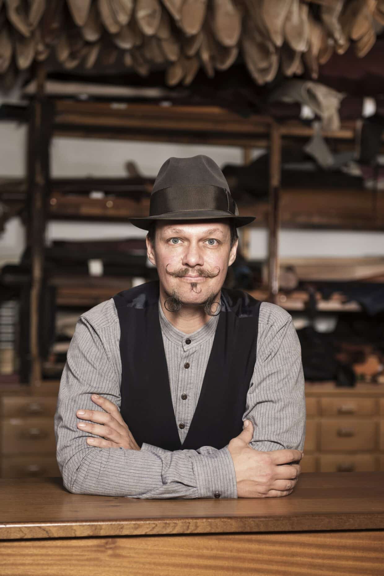Portrait_neu - Deutsche Manufakturenstrasse Mann mit Hut und Schnurrbart, lächelt am Tisch, umgeben von Schuhmaterialien im Hintergrund.