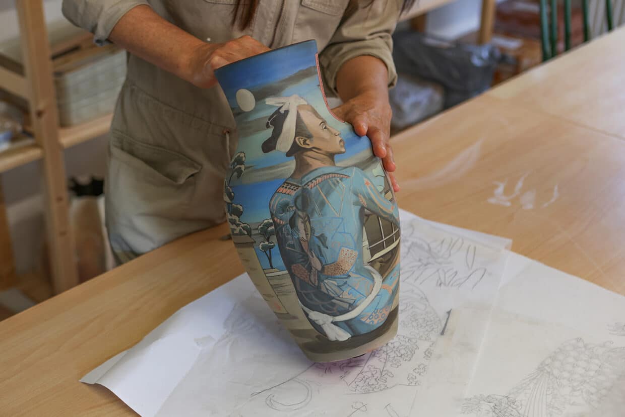 4_Victoria_Tobostai2025_074 - Deutsche Manufakturenstrasse Vase mit farbenfrohem, detailliertem Design, gehalten von einer Person in einem Atelier. Zeichnungen liegen auf dem Tisch.