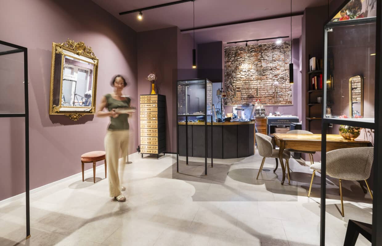 5082+_Ladengeschaeft_innen_Jun25_foto eberle - Deutsche Manufakturenstrasse Moderne Schmuckboutique mit einer Frau, die an einem Tisch vorbeigeht, und einer Backsteinwand im Hintergrund.