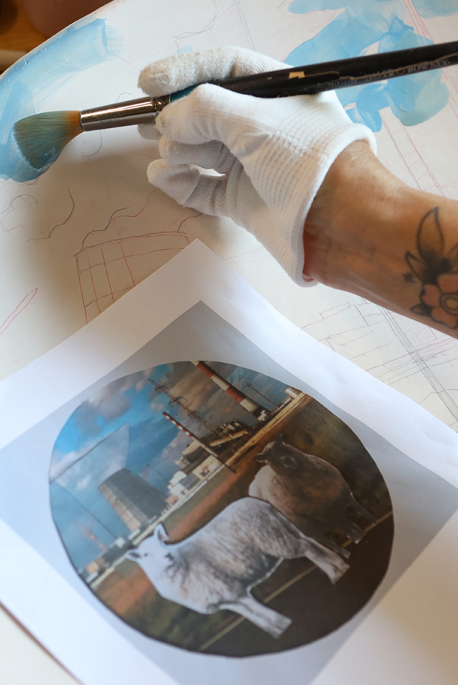 7_Victoria_Tobostai2025_063 - Deutsche Manufakturenstrasse Hand mit Pinsel in blauer Farbe, die auf eine Zeichnung auf Papier malt, daneben ein ausgedrucktes Bild eines Schafes und ...