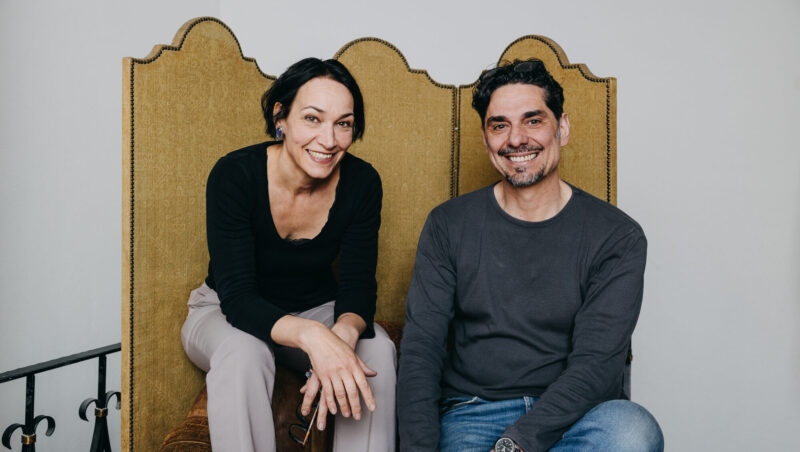 Silke Knetsch und Christian Streit sitzen lächelnd auf einer Couch mit einem goldfarbenen Rücken.