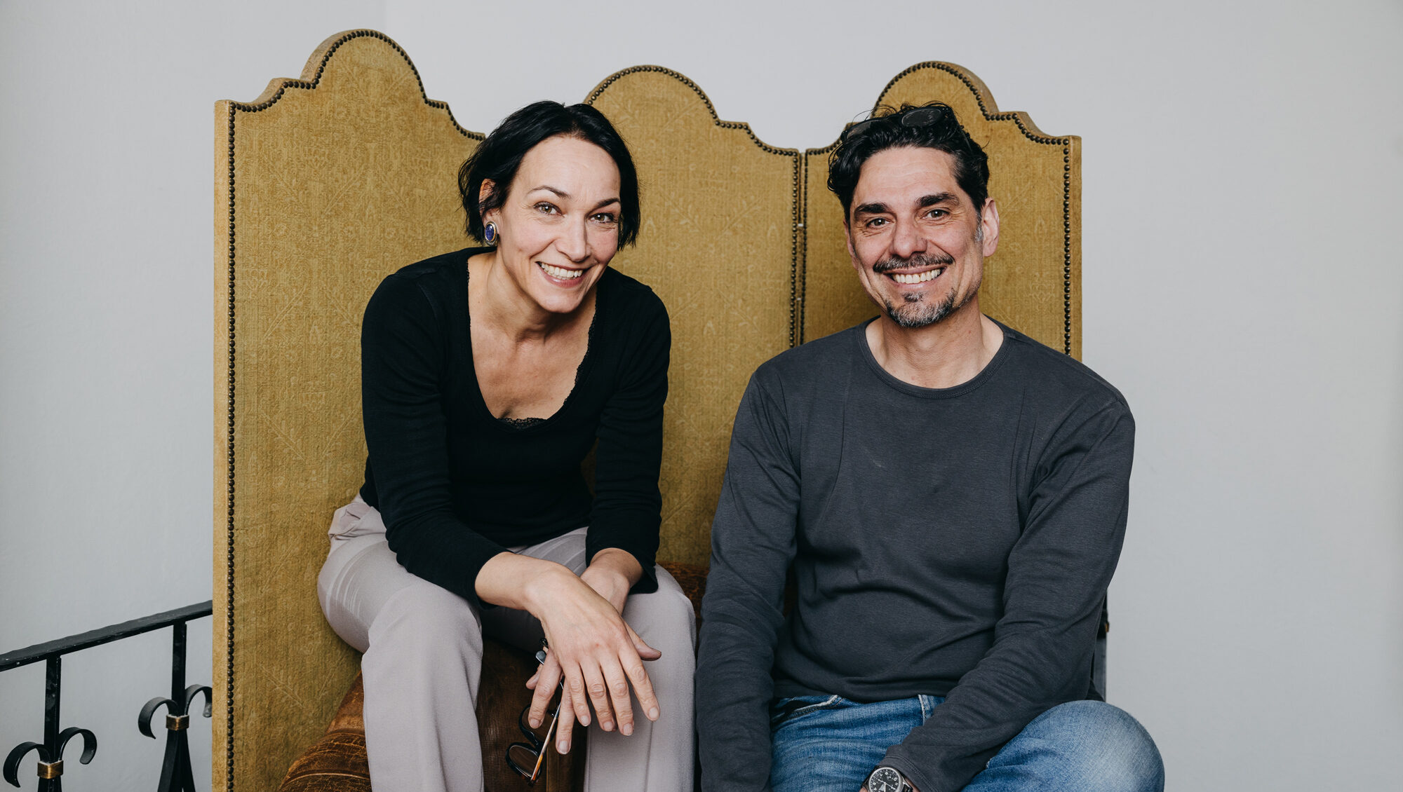 AG7A1251-groß - Deutsche Manufakturenstrasse Silke Knetsch und Christian Streit sitzen lächelnd auf einer Couch mit einem goldfarbenen Rücken.