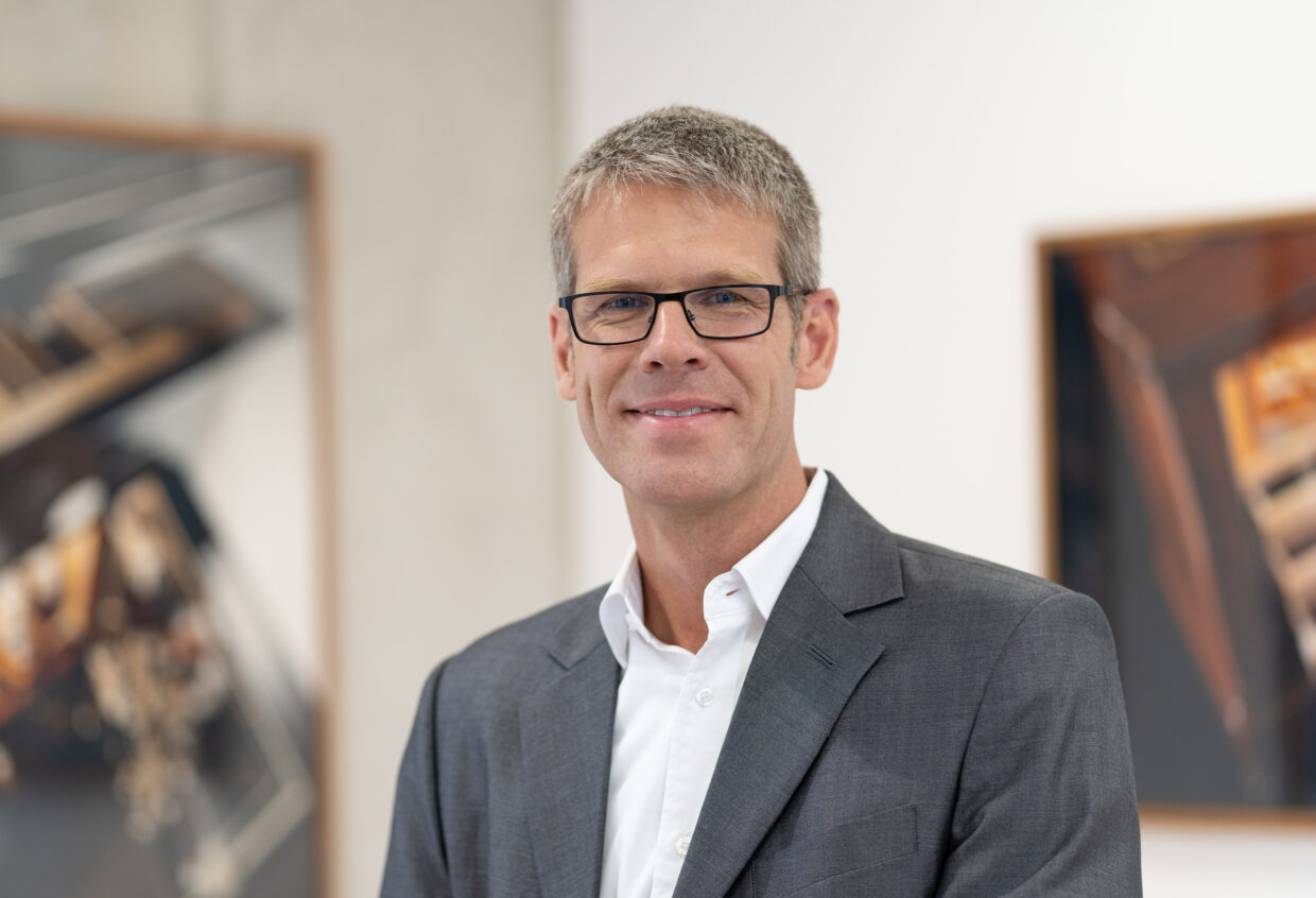 DOETTLING_SH_III - Deutsche Manufakturenstrasse Mann in Anzug mit Brille lächelt vor Kunstwerken in einem modernen Ausstellungsraum.