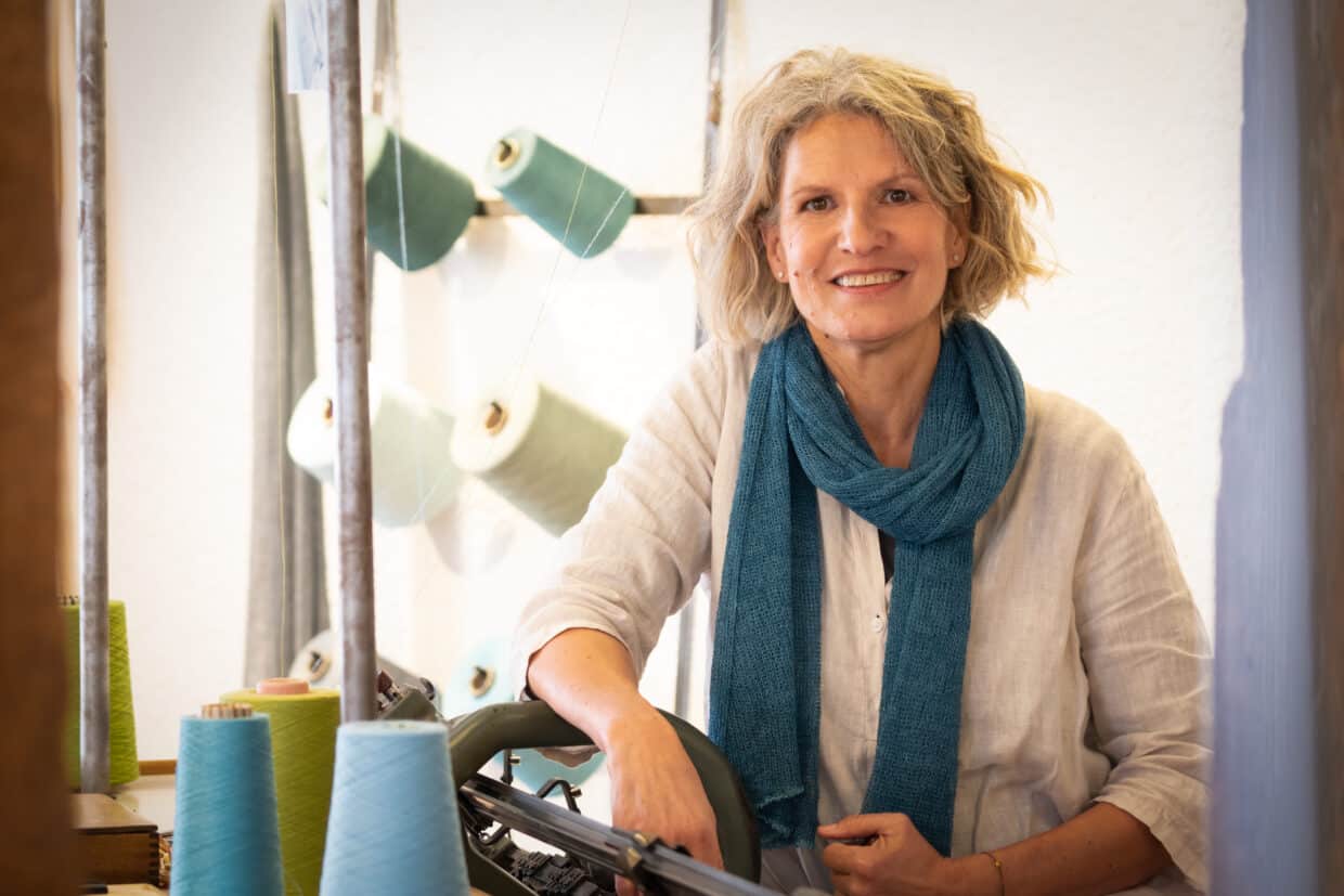 Grieb Uschi 2jpeg - Deutsche Manufakturenstrasse Uschi Grieb lächelt in ihrer Werkstatt, umgeben von bunten Garnrollen und einer Strickmaschine.