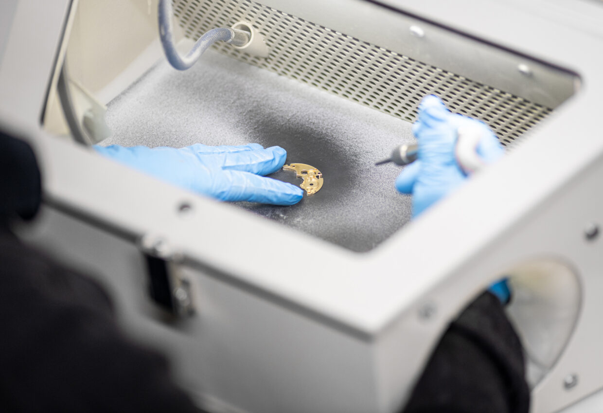 Lehmann-Sandstrahl-RGB - Deutsche Manufakturenstrasse Hand mit blauen Handschuhen bearbeitet ein goldenes Bauteil in einer Sandstrahlkabine.