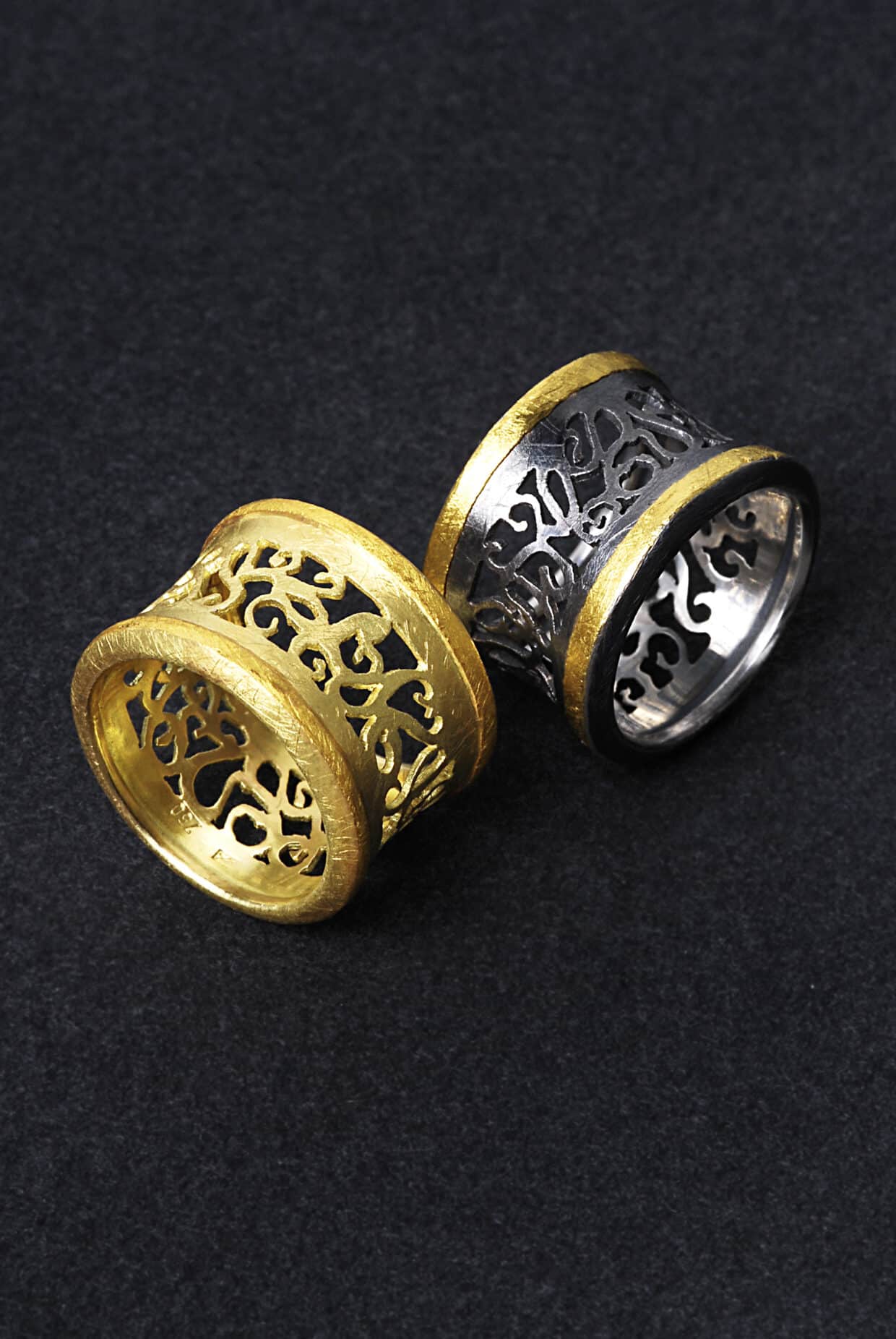 Ring_05 - Deutsche Manufakturenstrasse Zwei kunstvoll gestaltete Ringe, einer in Gold mit filigranem Muster, der andere in Schwarz und Silber.
