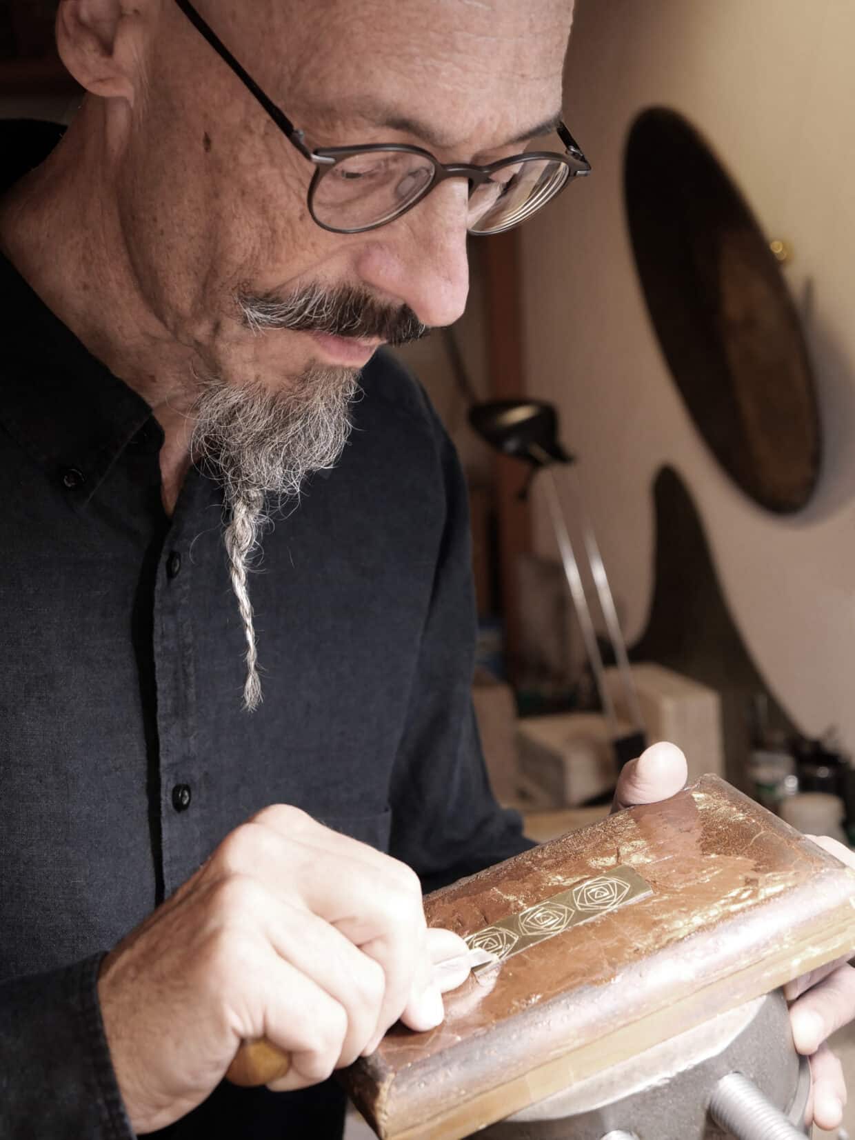 Tobias Dingler_ Gravur_sepia - Deutsche Manufakturenstrasse Mann mit Brille graviert eine goldene Fläche mit floralen Motiven in einer Werkstatt.