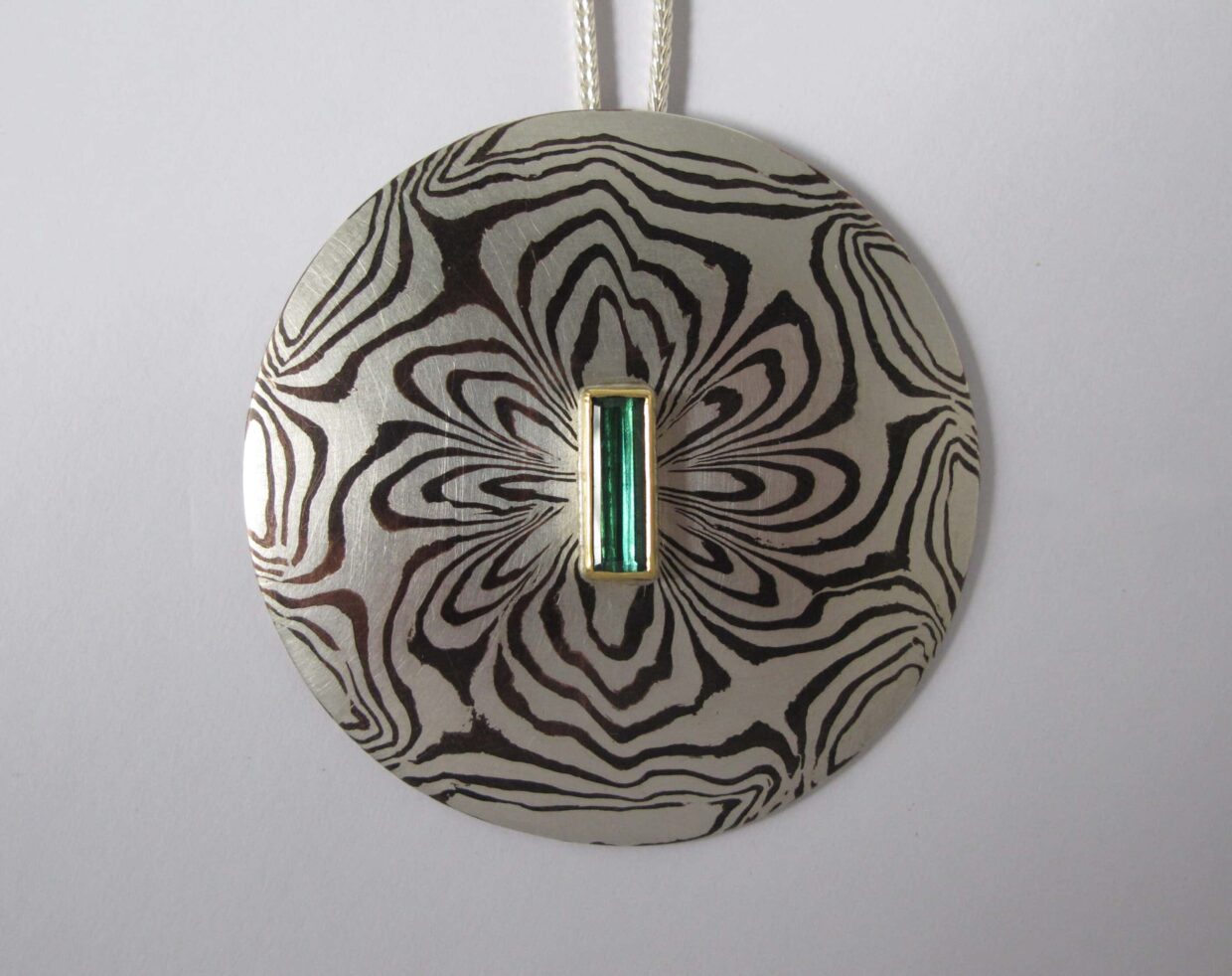 Tobias Dingler_Collier 1803 - Deutsche Manufakturenstrasse Runder Anhänger aus Metall mit einem zentralen, rechteckigen, grünen Edelstein und einem floralen Muster.