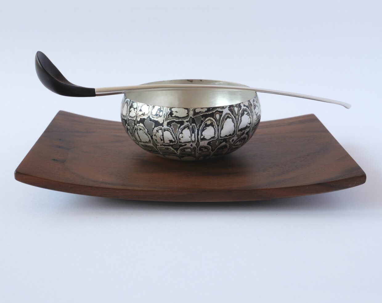Tobias Dingler_Kräuterschale_2 - Deutsche Manufakturenstrasse Kräuterschale aus Metall mit dekorativem Muster auf einer Holzplatte, daneben ein Löffel mit schwarzem Griff.