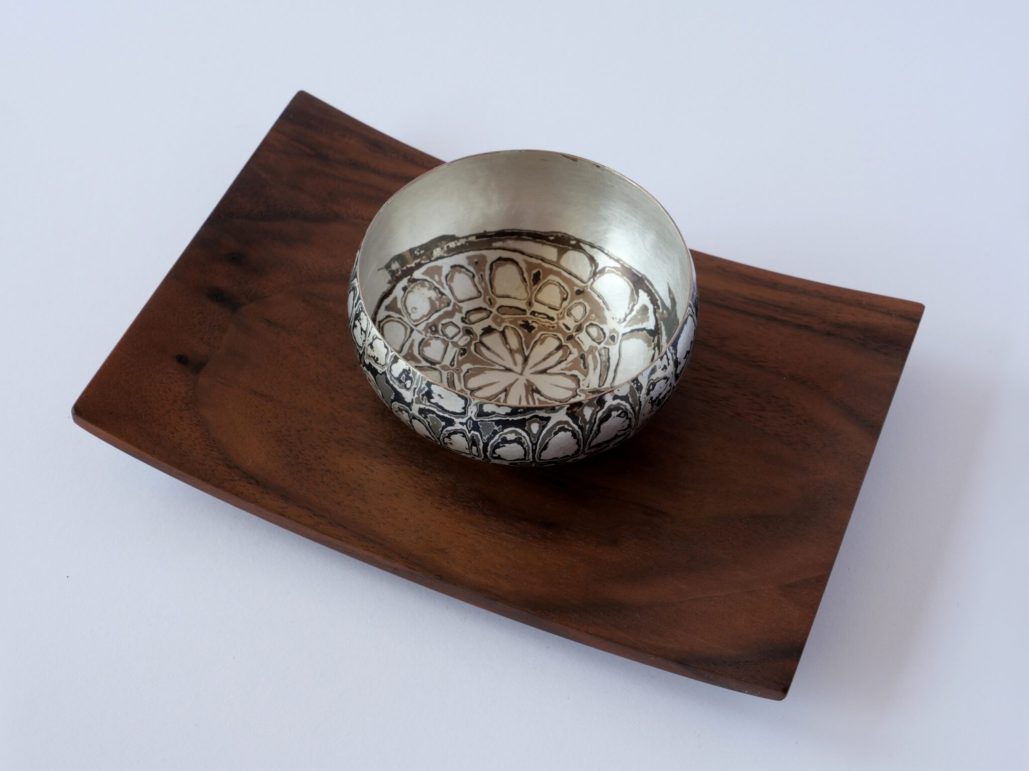 Tobias Dingler_Kräuterschale_3 - Deutsche Manufakturenstrasse Silberne Kräuterschale mit floralem Muster auf einer rechteckigen Holzplatte.