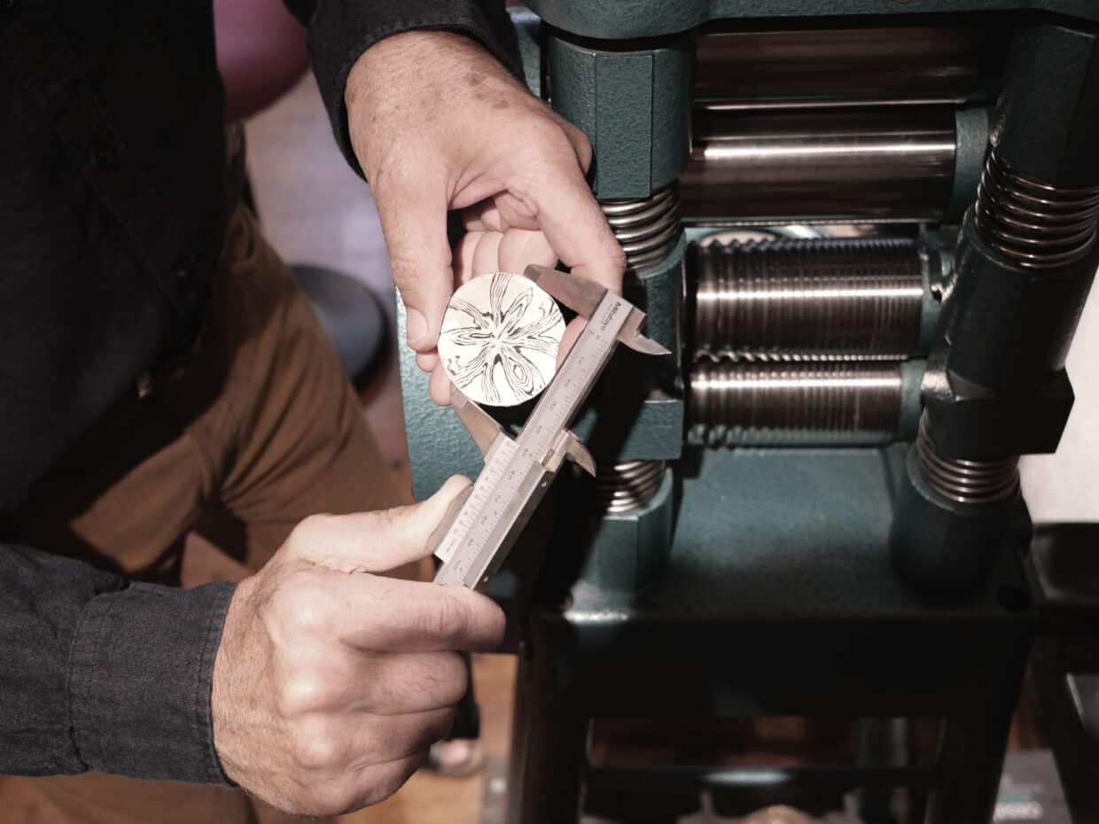 Tobias Dingler_Maß nehmen_sepia - Deutsche Manufakturenstrasse Hände messen einen runden, verzierten Metallgegenstand mit einem Messschieber an einer Maschine.