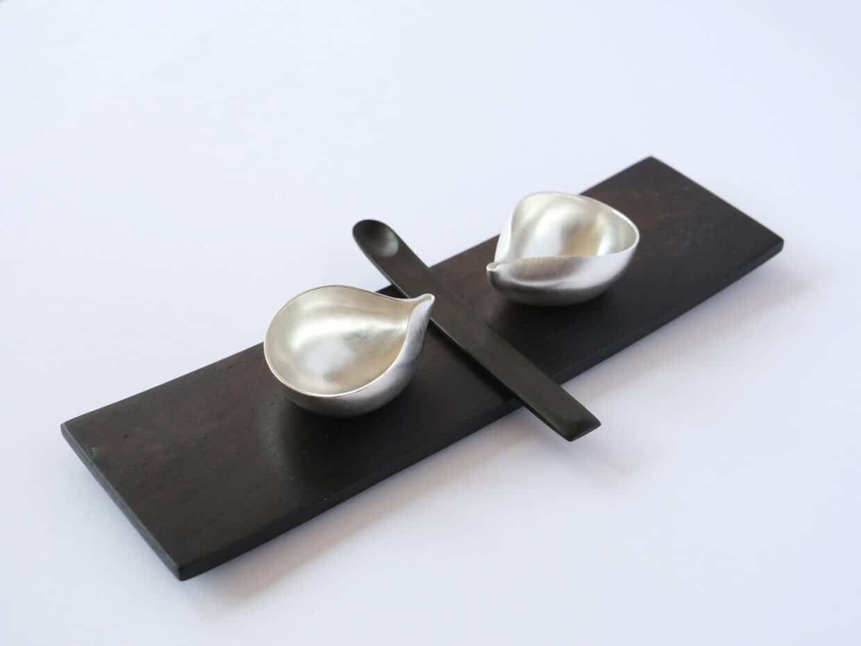 Tobias Dingler_Salz und Gewürz - Deutsche Manufakturenstrasse Zwei silberne Schalen und ein schwarzer Löffel auf einem rechteckigen Holzbrett.