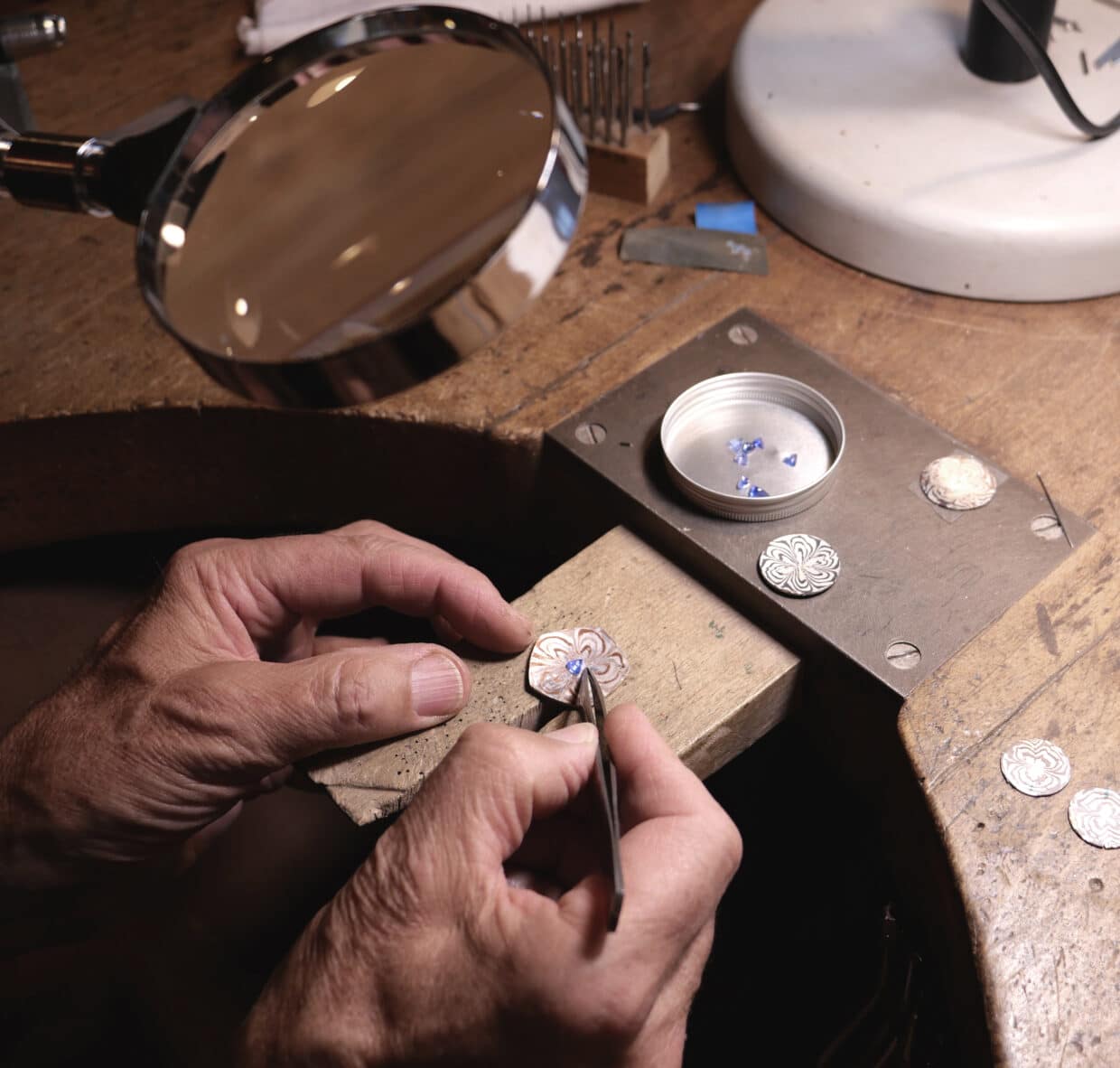 Tobias Dingler_Werkbrett_sepia - Deutsche Manufakturenstrasse Hände arbeiten an einem verzierten Metallstück auf einem Werkbrett, umgeben von Werkzeugen und einer Lupe.