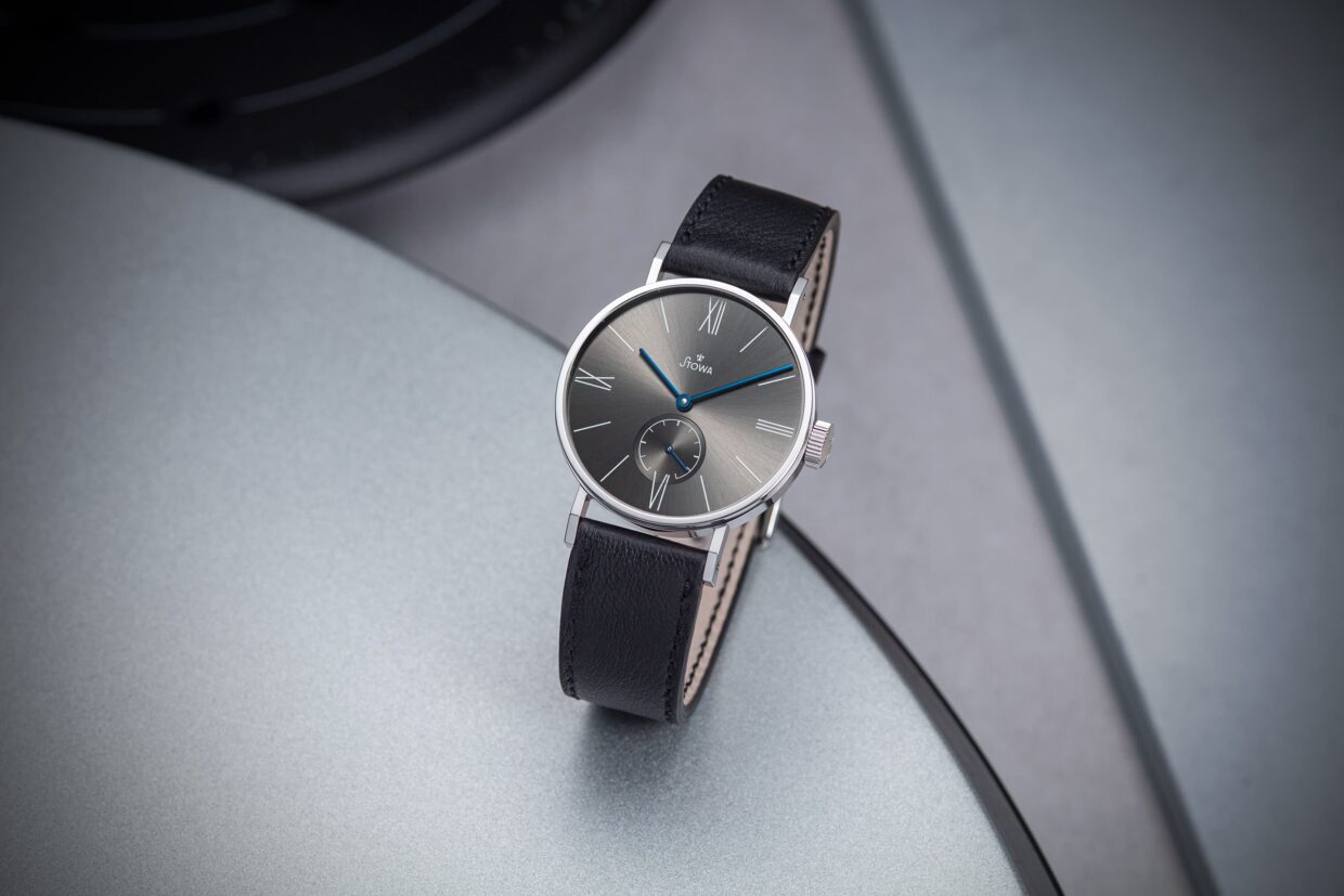 Elegante graue Armbanduhr mit schwarzem Lederarmband, römischen Ziffern und blauen Akzenten auf einem silbernen Untergrund.