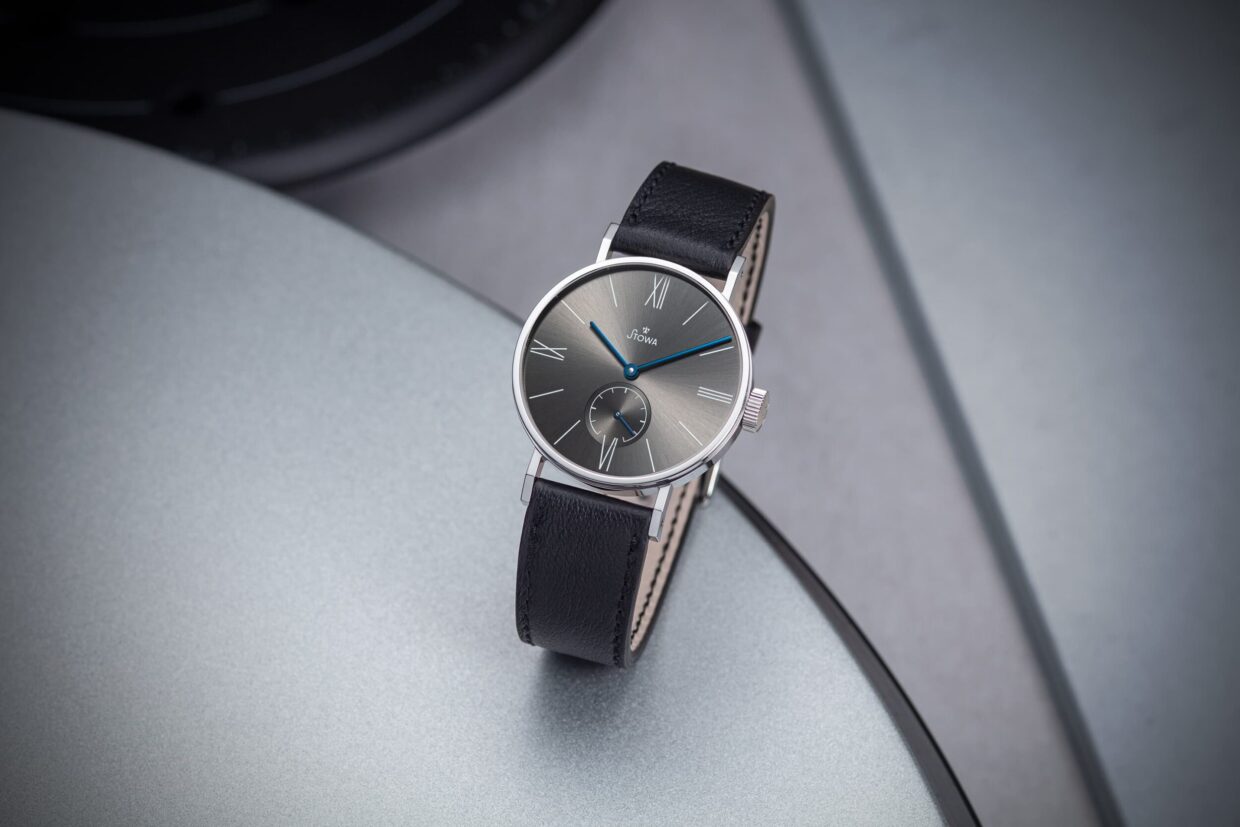 Antea-Klassik-KS-grau-01-scaled - Deutsche Manufakturenstrasse Elegante Armbanduhr mit grauem Zifferblatt, römischen Ziffern und schwarzem Lederarmband, auf einer silbernen Oberfläche...