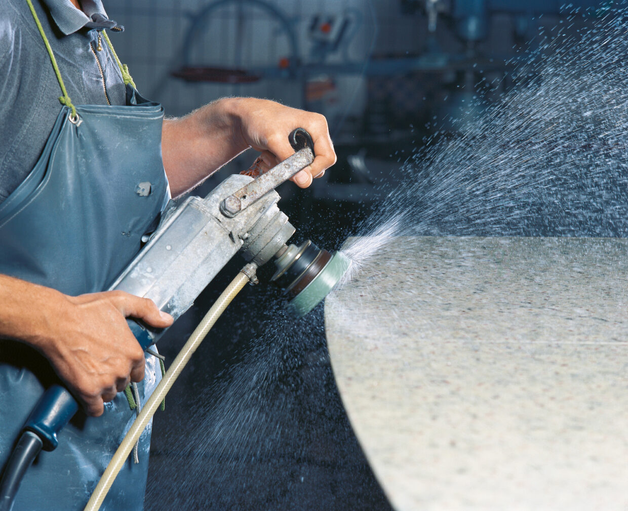 DRAENERT_Steinschleifen1 - Deutsche Manufakturenstrasse Arbeiter schleift eine Steinoberfläche mit einer Maschine, Wasser spritzt dabei auf die Fläche.